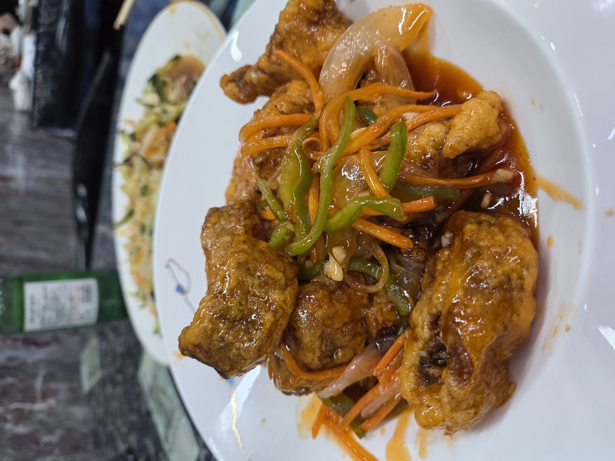 A close-up, top-down shot showcases a white oval dish filled with a vibrant, appetizing meal. The main subject is a sweet and sour dish, likely battered meat or fish, generously coated in a glossy, reddish-orange sauce. The protein pieces are deep-fried to a golden-brown hue. Adorning the dish are fresh, julienned orange carrots, sliced green bell peppers, and translucent white onions, adding color and texture. Small, lighter-colored pieces, possibly minced garlic or ginger, are visible within the rich sauce.
In the blurred background, another white plate is partially visible, containing what appears to be fried rice or noodles mixed with vegetables, suggesting a larger meal with multiple dishes. The plates rest on a dark, reflective surface, likely a dining table. To the left of the main dish, a blurry green bottle with an indistinct white label can be seen, hinting at a beverage accompanying the meal. The setting appears to be indoors, possibly a restaurant or home dining area, under artificial lighting. The focus is entirely on the food, presented as freshly prepared and ready to be enjoyed. No people are visible, and no specific time of day or location can be determined beyond an indoor dining context. There is illegible text on the green bottle in the background.