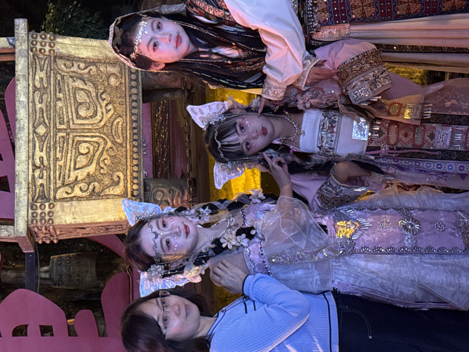 The image depicts four individuals posing in an artificially lit environment within Yuzhong District, China.
From left to right:
An adult woman, with short dark hair and eyeglasses, wears a light blue long-sleeved top with a black stripe at the hem and dark trousers. Her right hand rests on the left shoulder of the adjacent individual. She is looking towards the viewer with a slight smile.
Next to her stands a young woman in an elaborate light purple and white costume featuring sheer overlays, silver embellishments, and white floral details. She has large, translucent, butterfly-shaped headwear, a silver headband with a crescent moon motif, and multiple pearl necklaces. Her face is made up with white glitter patterns, red eyeshadow, and pink lipstick. Her dark hair is styled in braids adorned with flowers and beads. Her left hand gently touches the right arm of the person to her right. She gazes slightly to her left with a faint smile.
Adjacent is a younger woman dressed in a pink and white costume, including a white bandeau top with patterned trim and a pearl necklace. She wears similar butterfly-shaped headwear. Her face has glitter markings, prominent blush, and pink lipstick, with dark hair and bangs. Her left hand is near her own ear, while her right hand touches the left ear/hair of the individual beside her. She looks directly at the viewer with a slightly pouting, neutral expression.
On the far right, a young woman is depicted in a dark, intricately patterned costume with red and gold embroidery, a cream-colored sleeve, and a metallic headband. Her dark hair is styled with braids, beads, and loose strands. Her face features glitter markings and red lips. She looks directly at the viewer with a serious expression. Her left arm is partially visible, with her hand positioned on the third woman's side.
The background features a large, ornate, gold-colored structure, possibly a replica of an ancient Chinese ceremonial vessel, decorated with intricate patterns. To the left, a partial red character and decorative bells are visible. The scene is illuminated by artificial light, creating a warm glow against a dark, indistinct background. The overall environment suggests a cultural event or theatrical setting.