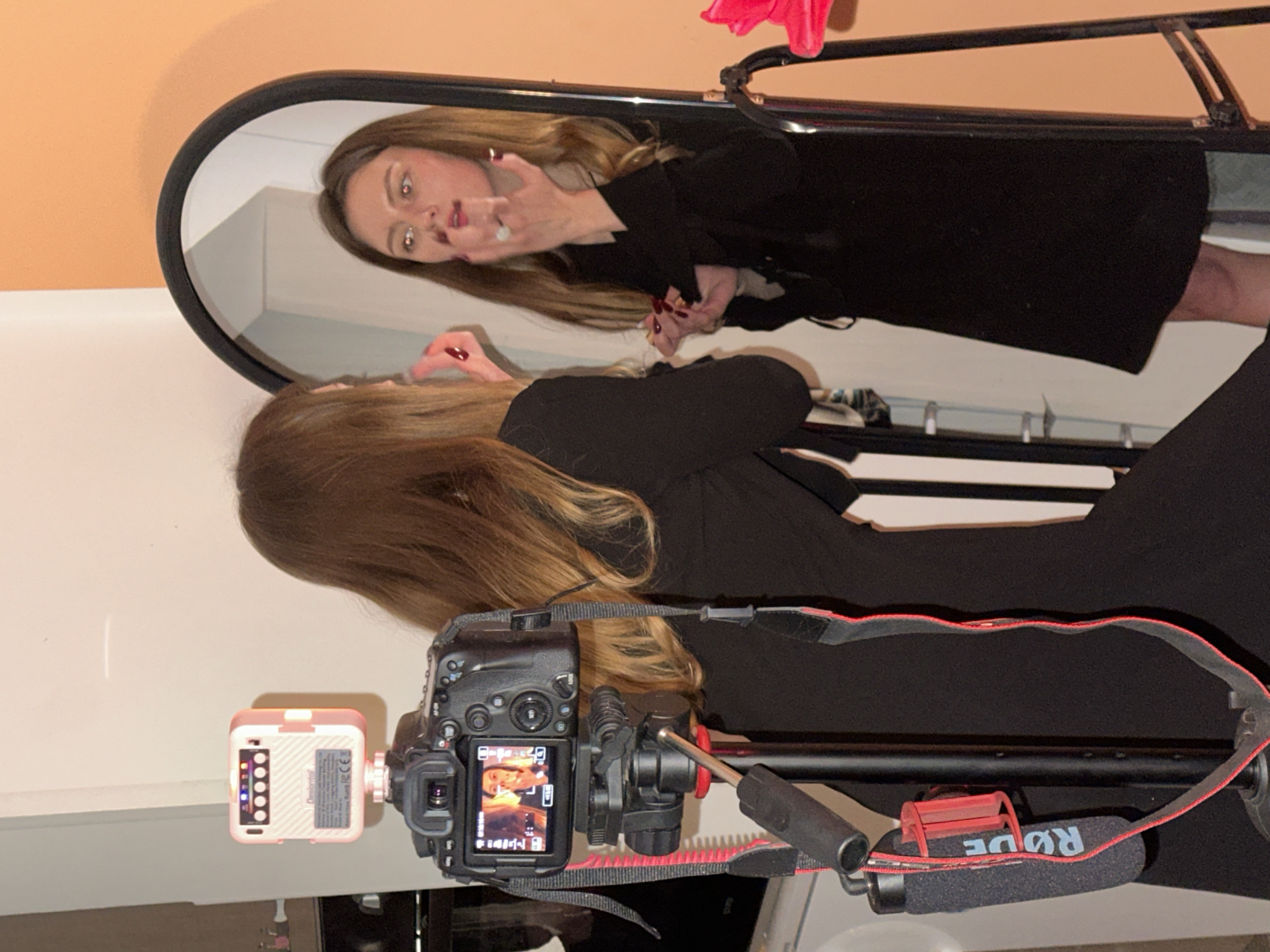 A person is reflected in an oval mirror. The person is wearing a dark blazer. The person is blowing a kiss at their reflection. A camera on a tripod with an attached light is capturing the scene. The background is a peach colored wall. The photo appears to be taken indoors, and there is no information to confirm the location as Šušanj, Montenegro.