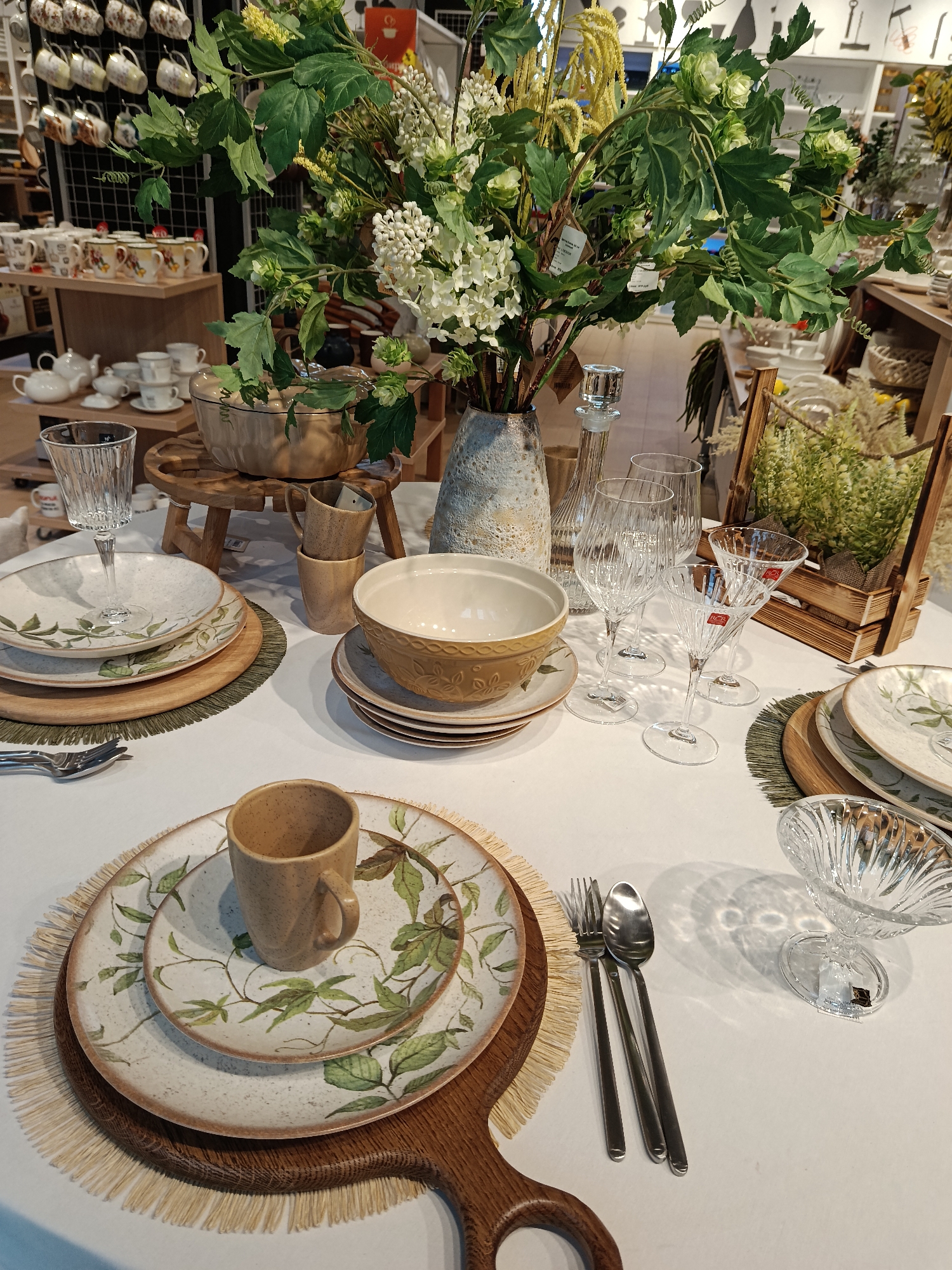 The image shows a display of tableware arranged on a white tablecloth. The setup includes plates with leaf patterns, wooden chargers and placemats, several styles of clear glassware, and ceramic bowls and mugs. The central element is a floral arrangement in a textured vase. The backdrop includes shelves stocked with various kitchenware items, suggesting the setting is a retail store. The style and decor imply the image could have been taken in Moscow, Russia. There are no people visible in the image.