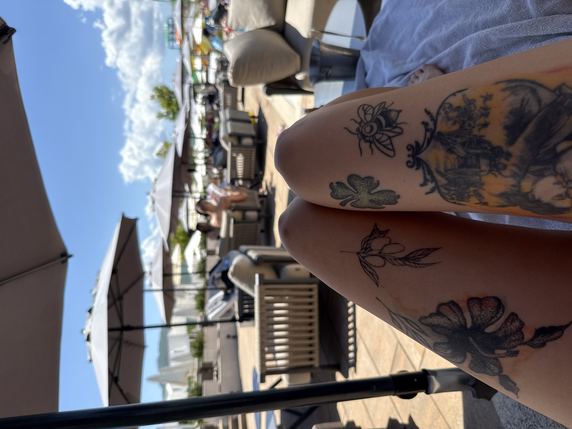 The image presents a first-person perspective, looking down over a person's tattooed legs towards an outdoor leisure area under bright sunlight. The legs, reclined on a light grey towel, display several distinct tattoos: on the left leg, a dark floral design and an olive branch; on the right leg, a four-leaf clover, a bee, and a large framed tattoo featuring a figure and a landscape. To the right of the legs, a silver ice bucket rests on a small, light-colored table.
Beyond the immediate foreground, a resort or hotel patio deck is visible, furnished with numerous outdoor lounge chairs and sofas with light-colored cushions, shaded by large grey patio umbrellas. Several indistinct individuals are present, seated and relaxing. The background features a clear blue sky with scattered white clouds, above distant, hazy hills or mountains. On the far left horizon, white structures are discernible, while colorful elements, suggestive of a play area or water park, are visible on the far right.