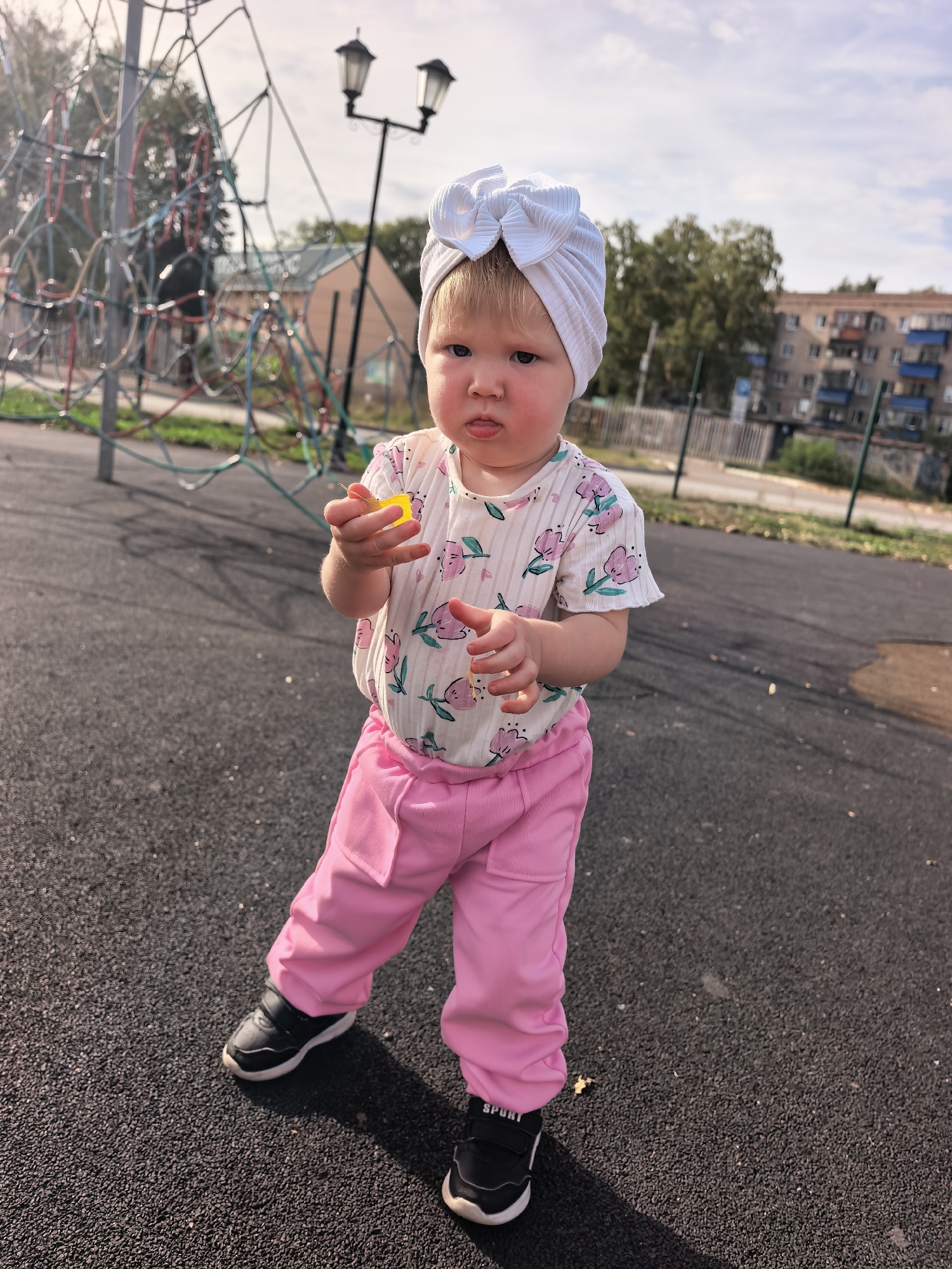 The image captures a young child standing on a dark, textured surface, likely a playground. The child is wearing a white turban with a large bow, a light-colored shirt with a floral pattern featuring pink flowers and green leaves, and bright pink pants. Black sneakers with white soles complete the outfit. The child's hands are visible, with the right hand holding a small yellow object. The background reveals a playground structure with colorful ropes, two lamp posts, trees, and residential buildings, suggesting an urban setting. The overall lighting indicates it's daytime, with some shadows visible on the ground. The scene is likely a public outdoor space in the city of Sterlitamak, Russia, given the architectural style of the buildings and the presence of a typical playground.
