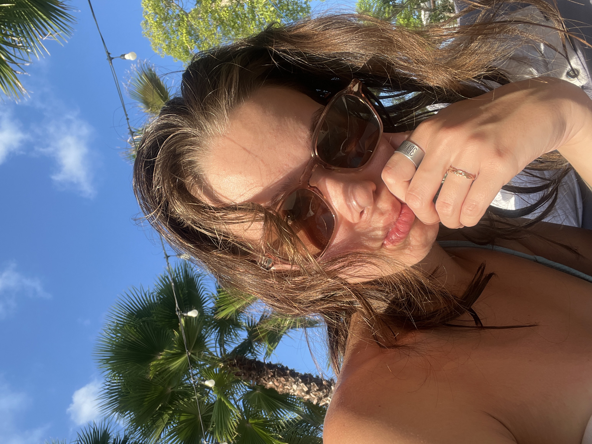 A low-angle outdoor selfie of a woman with brown hair and sunglasses. Her hand is near her face, displaying two rings on her fingers. The background includes a blue sky with some clouds, palm trees, and string lights. The location could be Nam Nha Trang Ward, Vietnam, but is not certain from the image alone.