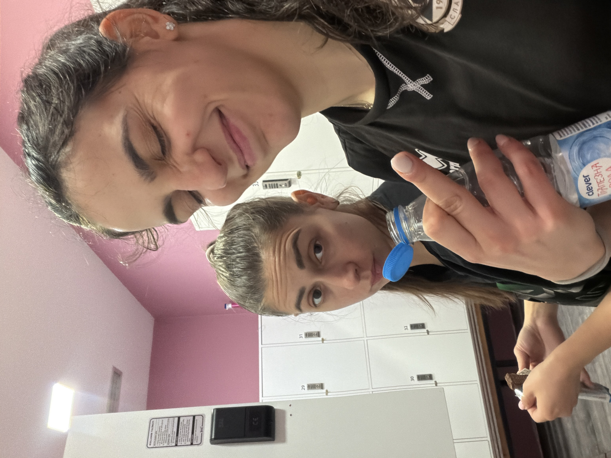Two young women are posing for a selfie in what appears to be a locker room. The room has white lockers with numbering (29-32 visible) against a pink and white wall. One woman is drinking from a "clever" brand water bottle, while the other is smiling at the camera. A chocolate bar is visible in the lower right corner. The city and country, Sofia, Bulgaria are presumed based on the water bottle label "clever ТРАПЕЗНА ВОДА", which translates to "clever TABLE WATER".