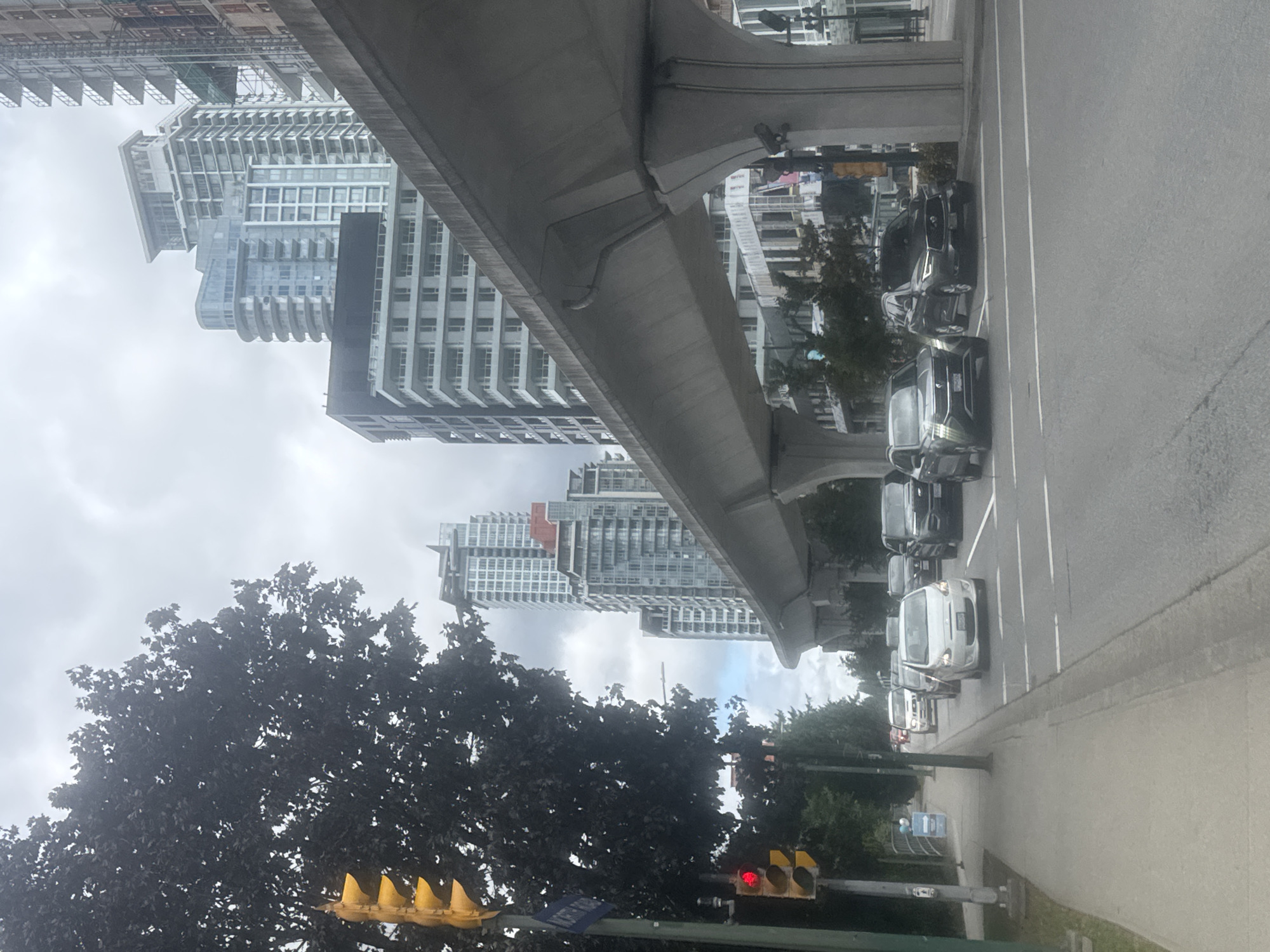 The image depicts a street scene in Coquitlam, Canada, with an elevated transit line running through the foreground. In the background, several modern high-rise buildings dominate the skyline under an overcast sky. Several vehicles, including cars and SUVs, are visible on the road, suggesting moderate traffic. A traffic light at the intersection shows a red light. Trees line the sidewalk on the left side of the street. The transit line's concrete structure is a prominent feature, supported by large pillars. The overall impression is of an urban environment with ongoing development and transportation infrastructure.
