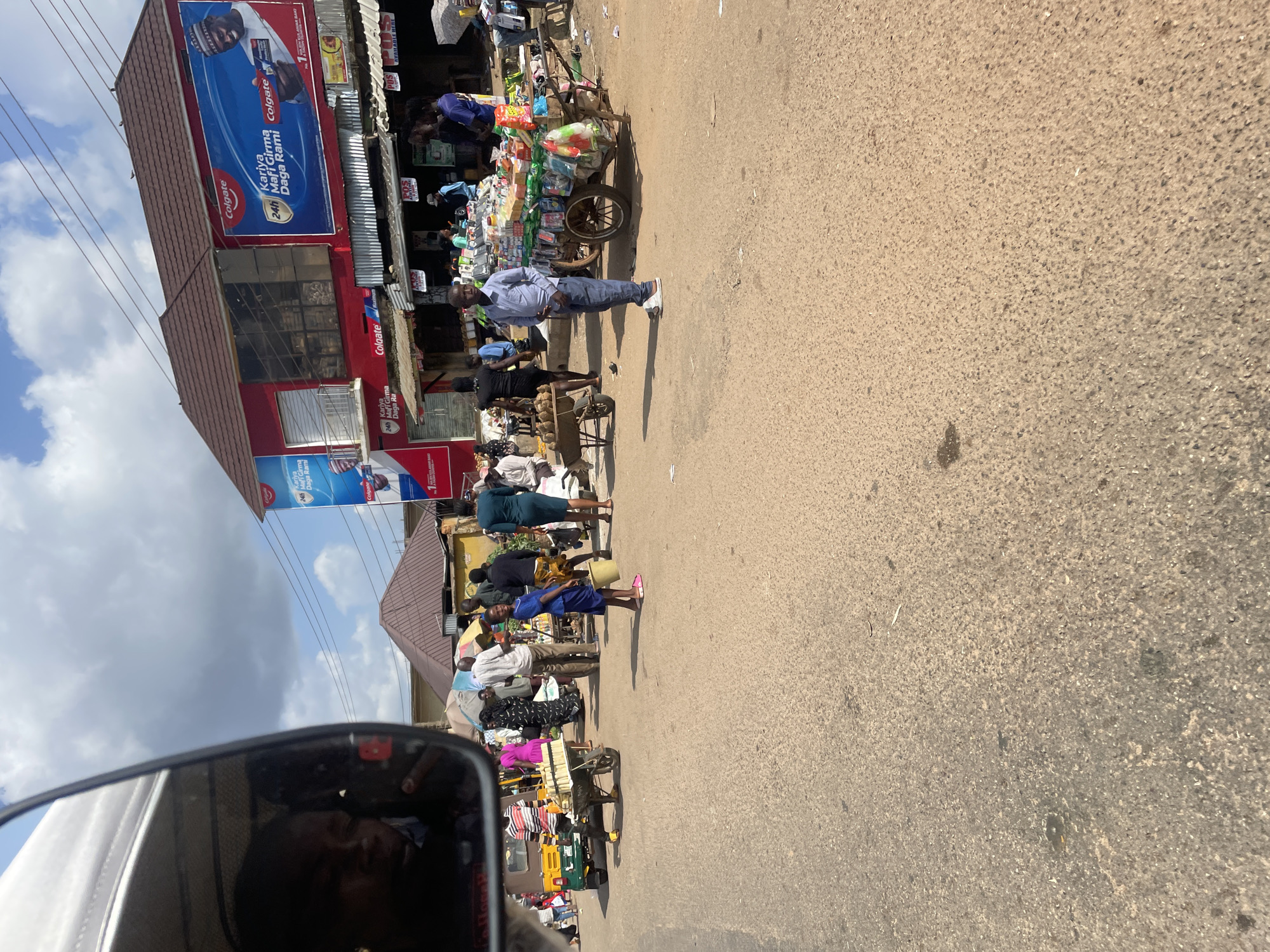 The image depicts a busy street scene, possibly a market, in an urban environment. In the foreground, a person is reflected in the side mirror of a vehicle, showing a child's face looking into the mirror. The street is paved with asphalt, and there are various market stalls and people engaged in activities.
Several people are visible in the midground, some standing and others walking. One person in the foreground is standing near a cart laden with goods, which appears to be a mobile stall selling various packaged items. Another cart, possibly carrying produce or other goods, is positioned further left. A young girl in a blue dress is walking with a yellow bucket.
In the background, a two-story building with a red facade and a brown roof is prominent. The building has large advertisements, including a Colgate advertisement featuring a man in traditional attire. Other market stalls and awnings are visible, suggesting a commercial area. The sky is partly cloudy. The overall impression is of a lively, everyday street activity in a town or city.
