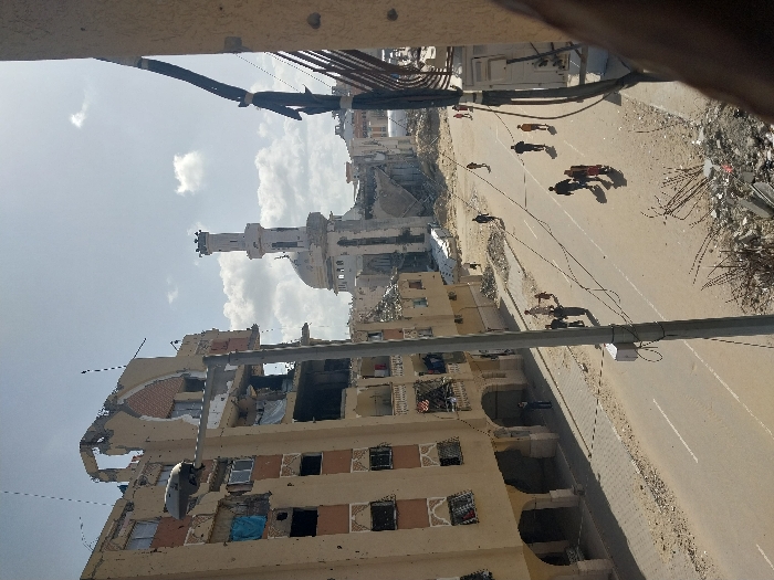 The image depicts a street scene in Khan Yunis, Palestinian Territory, characterized by extensive urban destruction under a partly cloudy sky. In the foreground to the left, a multi-story, light-tan residential building stands severely damaged, with upper floors partially collapsed, exposing interior rooms, rebar, and shattered windows. Blue tarpaulins are visible within some openings. The facade shows numerous impact marks.
A paved street, marked with two white lines, extends through the center of the frame into the distance. Along this street, multiple individuals, including adults and children, are seen walking in various directions. They appear to be civilians. Piles of rubble, debris, and twisted metal are scattered along the sides of the street. A utility pole with attached wires stands prominently in the mid-left.
In the background, a large mosque structure is visible, featuring a distinct minaret and a dome. The mosque also appears heavily damaged, with portions of its structure compromised and surrounded by debris. Further down the street, additional damaged buildings and piles of rubble are present. The right foreground shows the edge of another building and bundles of black cables. The overall scene conveys widespread destruction and civilian movement through a severely impacted urban environment.