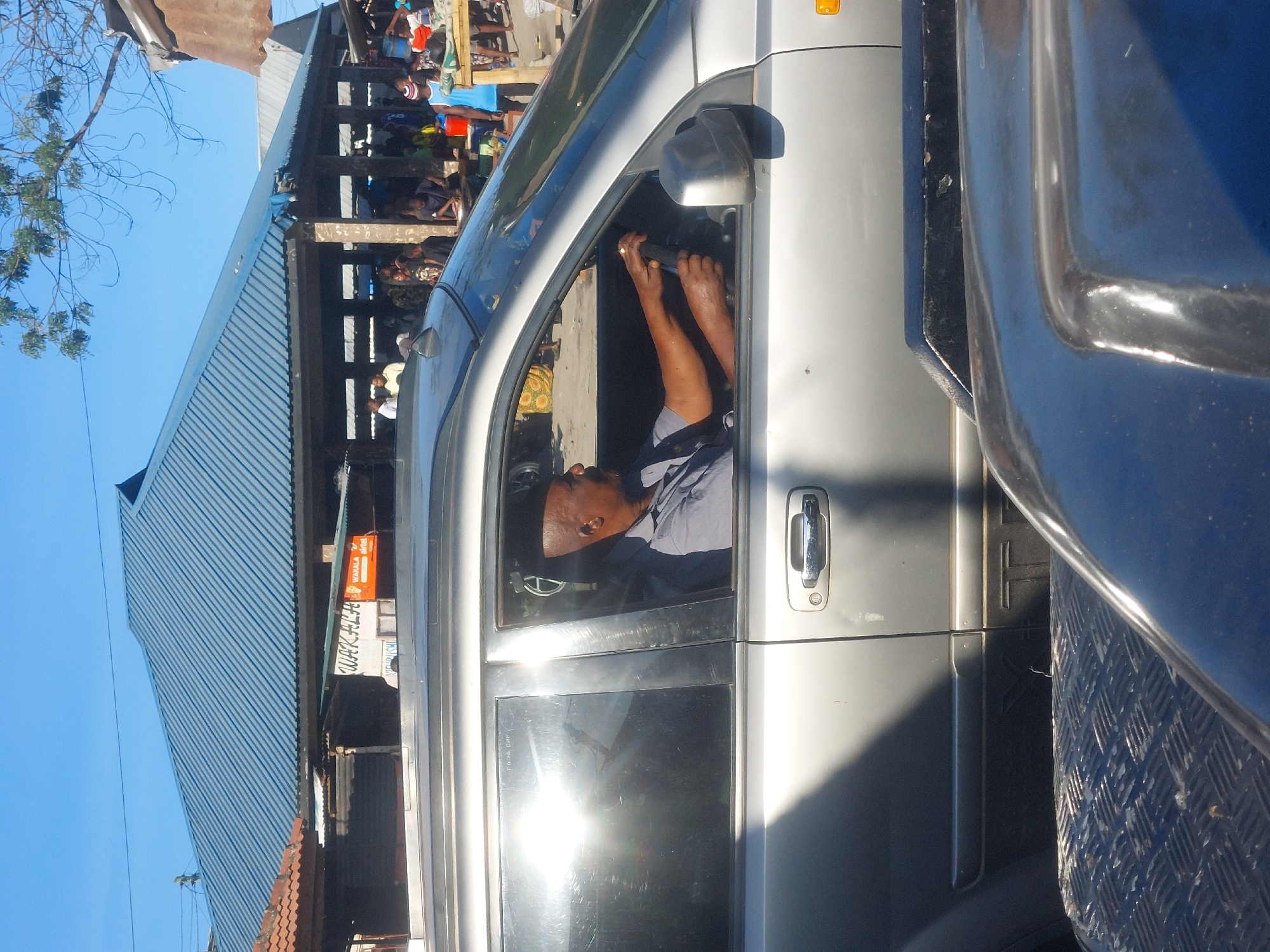 The image shows the interior of a silver van or SUV in what appears to be a busy urban area. A man is sitting in the driver's seat, holding the steering wheel with both hands. He is wearing a light-colored shirt, a dark blue undershirt and a dark cap. The sun is reflecting off the vehicle’s windows, obscuring details. Adjacent to the silver vehicle is a blue vehicle with a diamond-patterned surface. Behind the silver vehicle, there appears to be a covered walkway with a crowd of people. The background building has a corrugated metal roof. A sign reading "WAKALA airtel" is visible on the side of the building. This image was taken in Dar es Salaam, Tanzania.