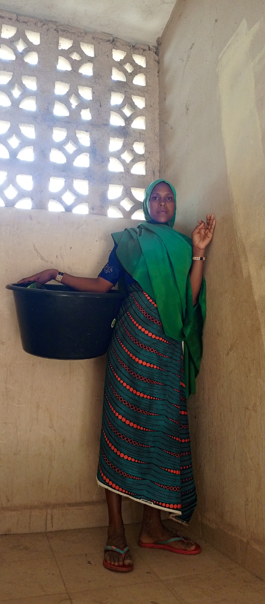 The image captures a woman standing indoors, likely within a structure with unpainted, textured earthen or mud-plastered walls. Bright daylight streams through a decorative ventilation wall on the left, creating illuminated patterns on the wall. The floor is tiled with light-colored squares.
The woman stands facing forward, with a neutral and serious expression. She is dressed in traditional attire, including a light green hijab covering her head and a matching green shawl draped over her left shoulder and arm. She wears a dark blue long-sleeved top and a long, patterned skirt featuring wavy rows of orange-red circles and dots on a dark teal background. A white border at the skirt's hem reveals faint black text, "VB18071202 SA". On her feet, she wears red flip-flops. She has a gold and black bracelet on her left wrist and a beaded bracelet on her right.
She holds a large, black, plastic basin or tub against her left hip, with her left hand resting inside it, suggesting it might contain water or items. Her right hand is gently placed against the textured wall to her right. The scene depicts a moment of daily life, likely during the daytime given the natural light source, in what appears to be a modest domestic setting.