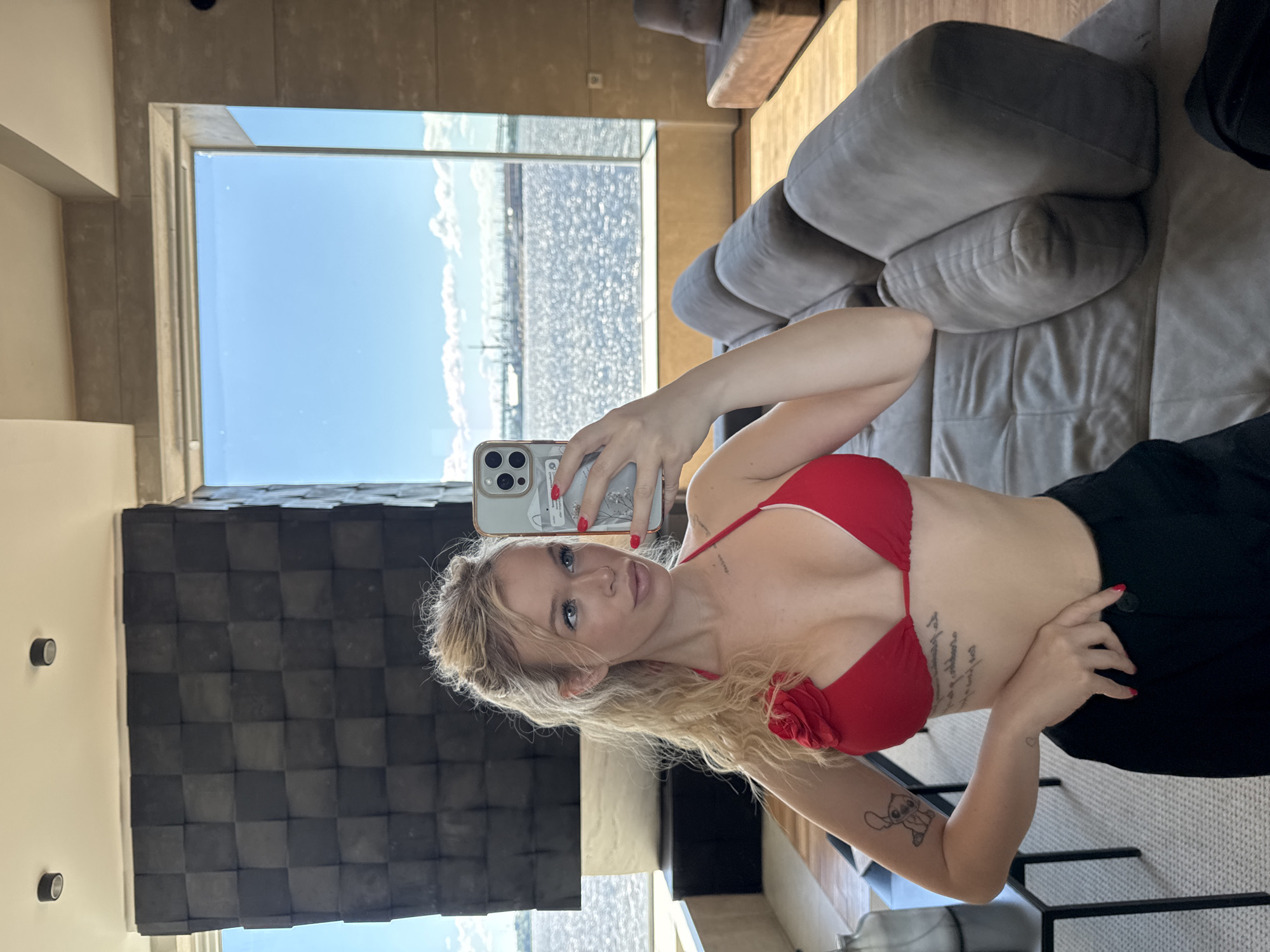 A young woman with blonde hair and blue eyes is taking a mirror selfie in a modern apartment overlooking the Gulf of Finland. She is wearing a red bikini top and black pants. The apartment features a large window with a view of the water and a cityscape, as well as a plush sectional sofa and contemporary decor. The woman's arm is visible, showing tattoos. The overall impression is one of luxury and relaxation.