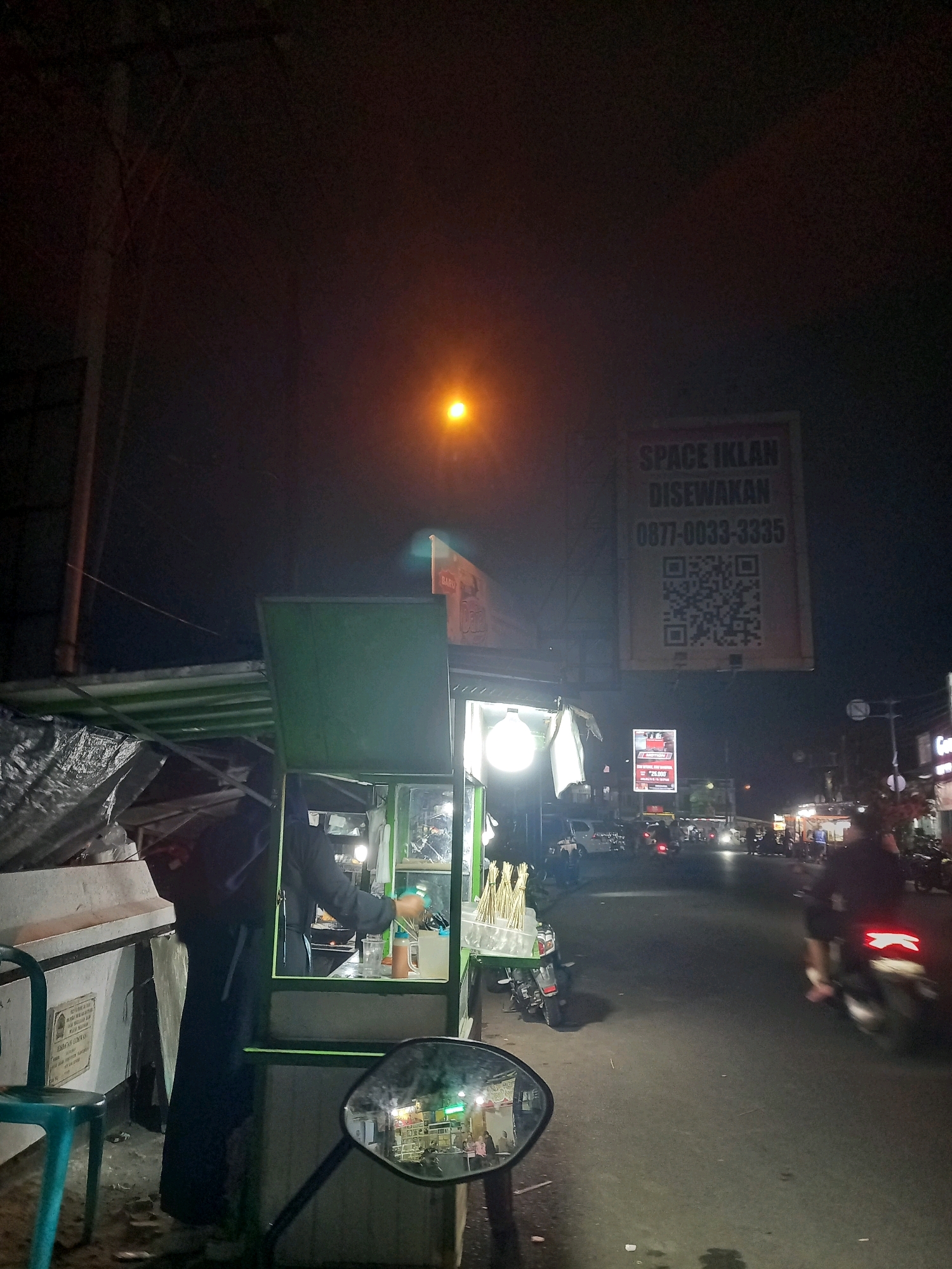 This is a low-light, nighttime, eye-level outdoor shot of a street scene in Bawang, Indonesia. The foreground shows a motorcycle's rearview mirror reflecting a busy street with various stalls and vehicles. To the left, a green food cart with an open service window is visible, with a person wearing dark clothing and a headscarf inside, seemingly preparing food. Next to the cart is a stack of what appear to be bundled items covered in a grey tarp and a turquoise chair. Above the food cart, a large, square advertisement sign reads "SPACE IKLAN DISEWAKAN" (Advertising Space for Rent) with a phone number "0877-0033-3335" and a QR code. The street itself is paved and appears to be a main thoroughfare with multiple lanes. Several motorcycles and a few cars are visible on the road, some with their headlights on. In the background, there are more dimly lit stalls and buildings, suggesting a commercial or market area. A bright streetlamp illuminates the upper right portion of the scene. The overall ambiance is that of a bustling night market or street food area.
