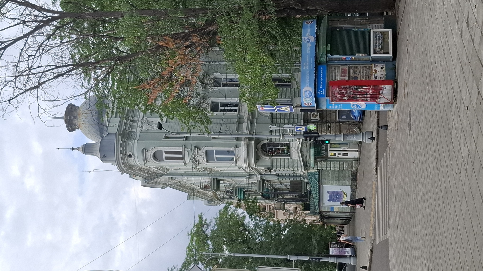 In an urban setting under an overcast sky, a light green, ornate building stands prominently on the right. The building features multiple floors with numerous windows and balconies, topped with a distinctive domed roof and spire. Adjacent to the building, on the sidewalk, is a blue kiosk with various products displayed. A large tree with green foliage partially obscures the building.
In the foreground, a paved pedestrian area with a crosswalk stretches across the street. Two individuals are visible crossing the street from left to right. A woman in a black dress carries a bag, while another woman in a white top and blue jeans walks ahead. Traffic lights indicate a green light. A street lamp stands on the left side of the image. The environment appears to be a city street with a mix of residential and commercial elements.