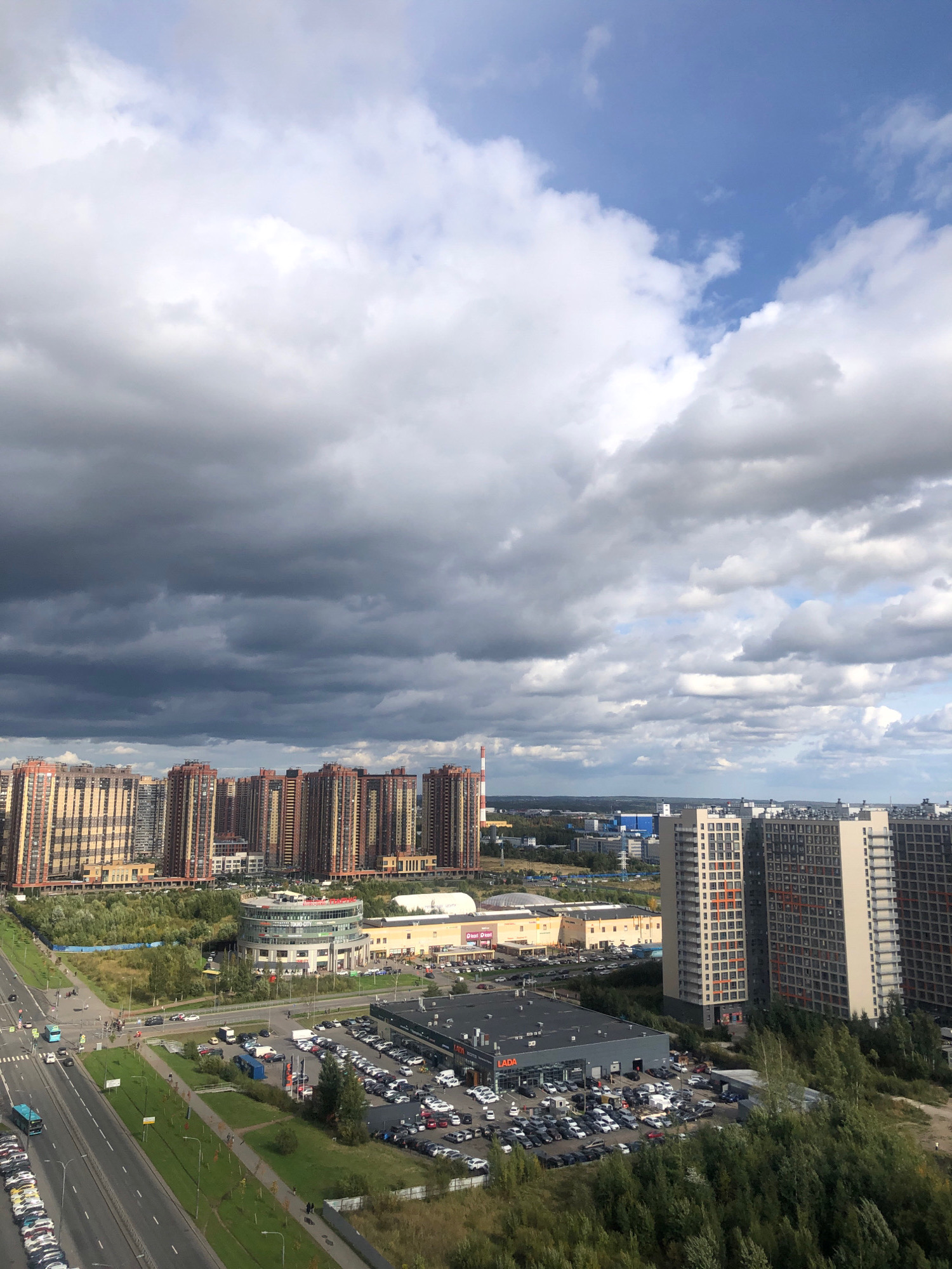 The image shows a panoramic view of the city of Vsevolozhsk, Russia, on a cloudy day. The foreground features a multi-lane road with several vehicles, including cars, buses, and trucks. Adjacent to the road are parking lots filled with cars, a Lada car dealership, and a shopping center with a circular building labeled "Eurograd." Behind these structures, numerous tall residential buildings with light-colored facades and orange window frames are visible. In the distance, more buildings, industrial facilities, and a smoking chimney are scattered across the landscape, with a distant horizon under a dramatic sky filled with dark, ominous clouds. The area is characterized by a mix of urban development and green spaces.