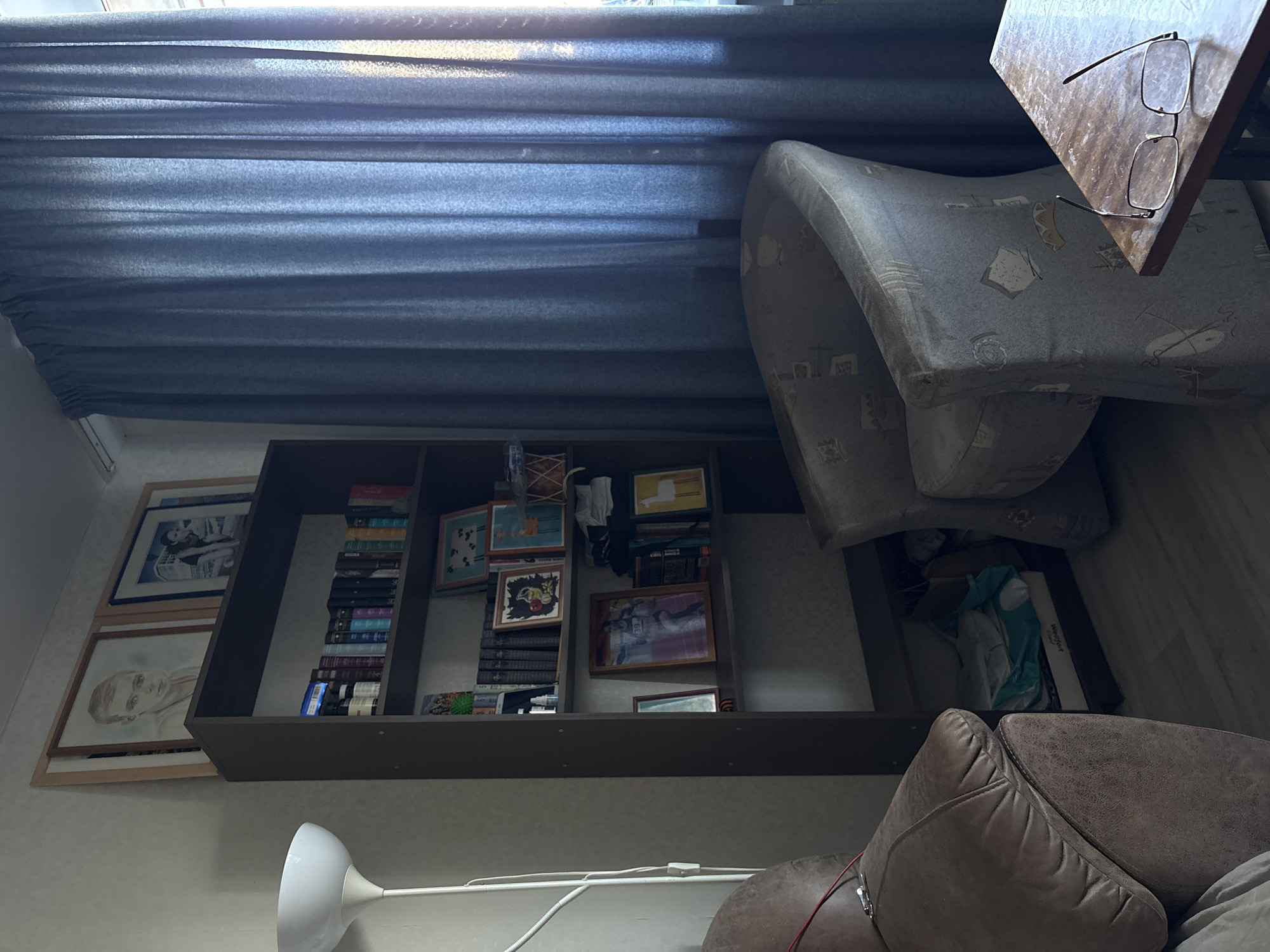 The scene is an interior shot of a room, likely in Ulyanovsk, Russia, given the prompt. A bookcase is positioned against a wall, filled with books, picture frames, and other personal items. Two framed pictures are hanging to the left of the bookcase. A lamp is visible near the bottom of the image. A worn armchair is on the right. A side table with glasses on it is next to the chair. Heavy blue curtains are drawn, providing soft light.