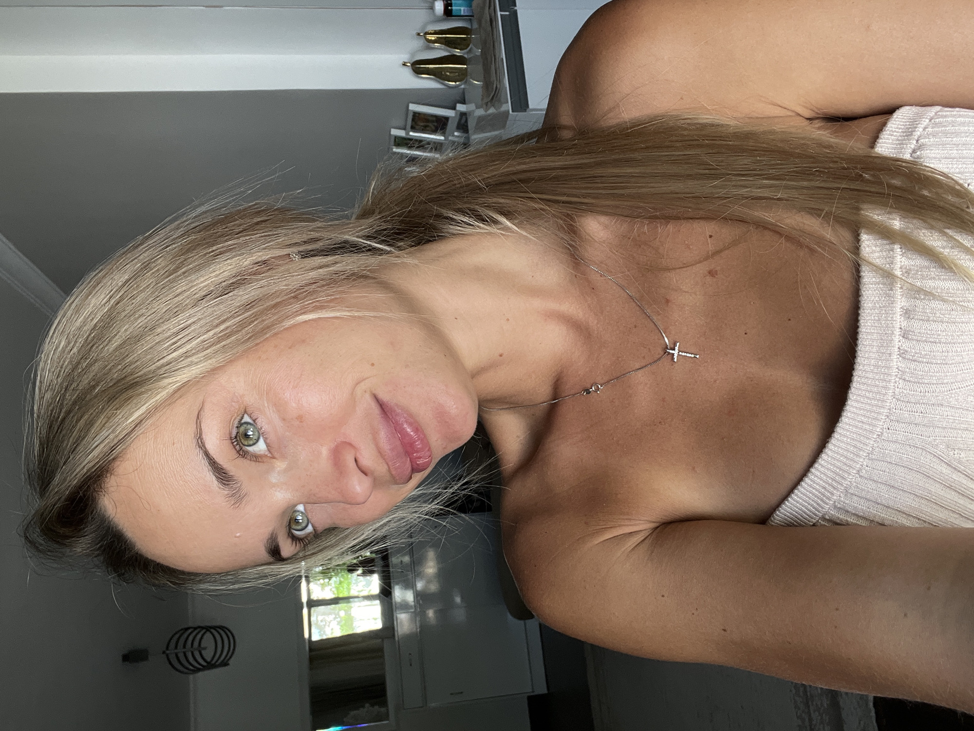 A close-up, waist-up portrait of a young woman with blonde hair tied in a ponytail, light skin with freckles, and green eyes. She is wearing a light-colored, strapless top and a silver necklace with a cross pendant. The background appears to be an indoor setting, possibly a room in Sharm El Sheikh, Egypt, with a window letting in natural light and some furniture visible. The lighting suggests it is daytime.