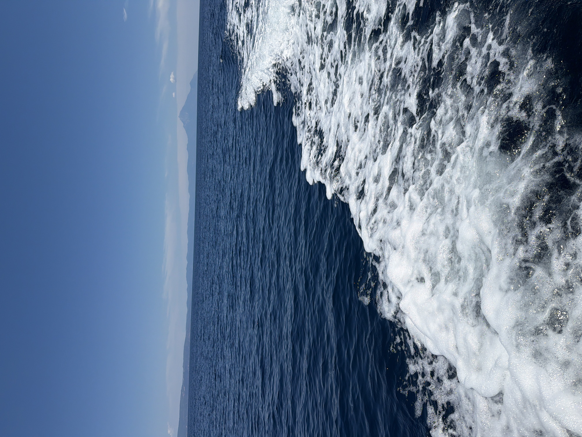 The image shows a view from a boat moving through the sea, creating a foamy wake on the right side of the frame. The water is a deep blue, and in the distance, a hazy mountain range is visible under a clear blue sky. On the horizon, a prominent pointed mountain, possibly Mount Athos, can be seen. The scene is likely a coastal area in Greece.