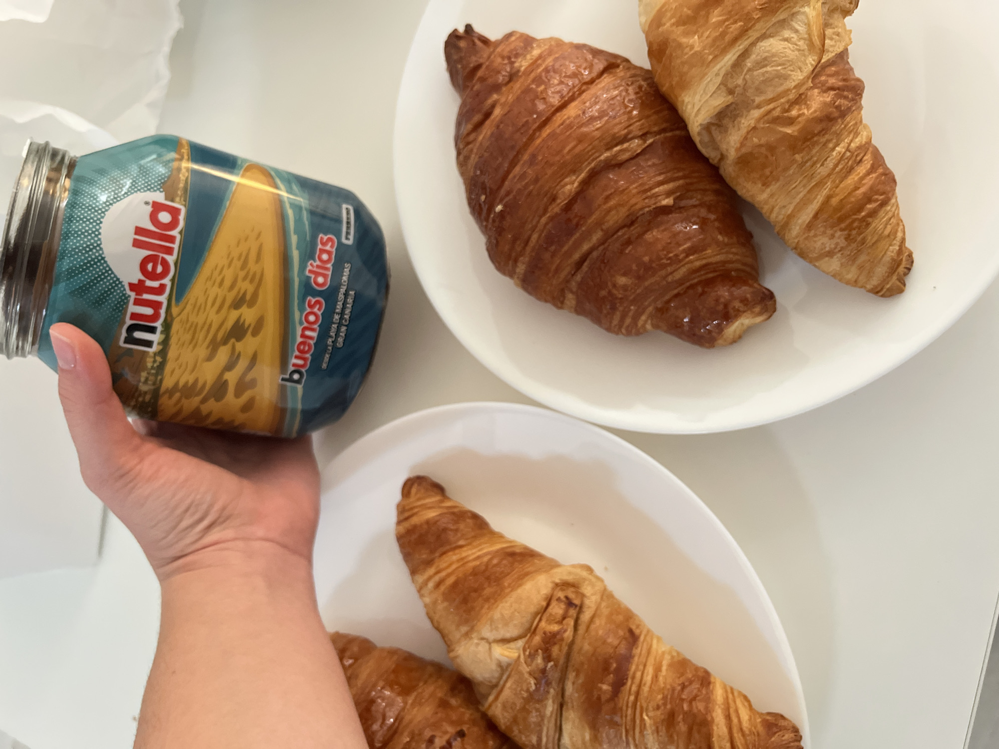 The scene depicts a person's left hand holding a jar of Nutella with a label that reads "Buenos Días" and indicates that the photo is taken "Desde la Playa de Maspalomas, Gran Canaria." Two white plates are on a white surface, each containing croissants. The top plate holds two croissants, one darker brown and more textured than the other. The bottom plate holds two croissants, both appearing lighter in color compared to the first croissant on the top plate.