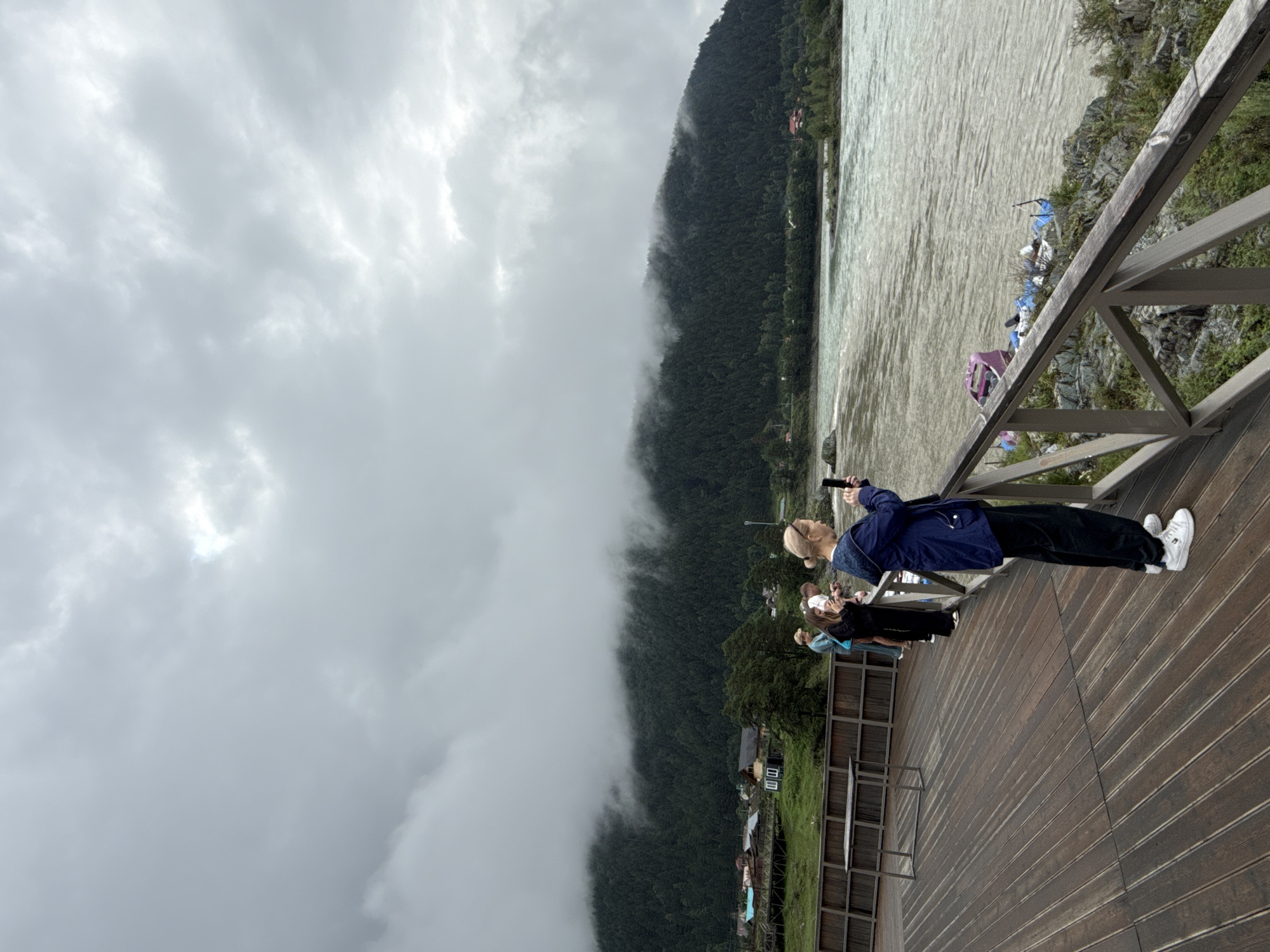 The image depicts a group of individuals standing on a wooden deck overlooking a wide, turbulent river. The sky is overcast with heavy clouds, and a dense forest covers the slopes of a mountain in the background. Some of the cloud cover appears to be hanging low on the mountain, obscuring its upper reaches.
In the foreground, several people are visible on the deck. A woman in a dark blue jacket and black pants is in the process of taking a photo with her phone, facing away from the camera. To her left, a man and two other individuals, one wearing a light blue top, are also looking out at the river. Further to the left, a metal table stands on the deck.
The river itself is murky and appears to have strong currents. Along the rocky shore on the far side of the river, there are several pieces of debris, including what appears to be a purple and pink structure, possibly part of a raft or boat, along with blue tarps and other assorted items. Small buildings and structures are visible nestled at the base of the forested mountain on the far bank. The overall scene suggests a natural, possibly remote, location in Elekmonar, Russia, under a brooding, cloudy sky.