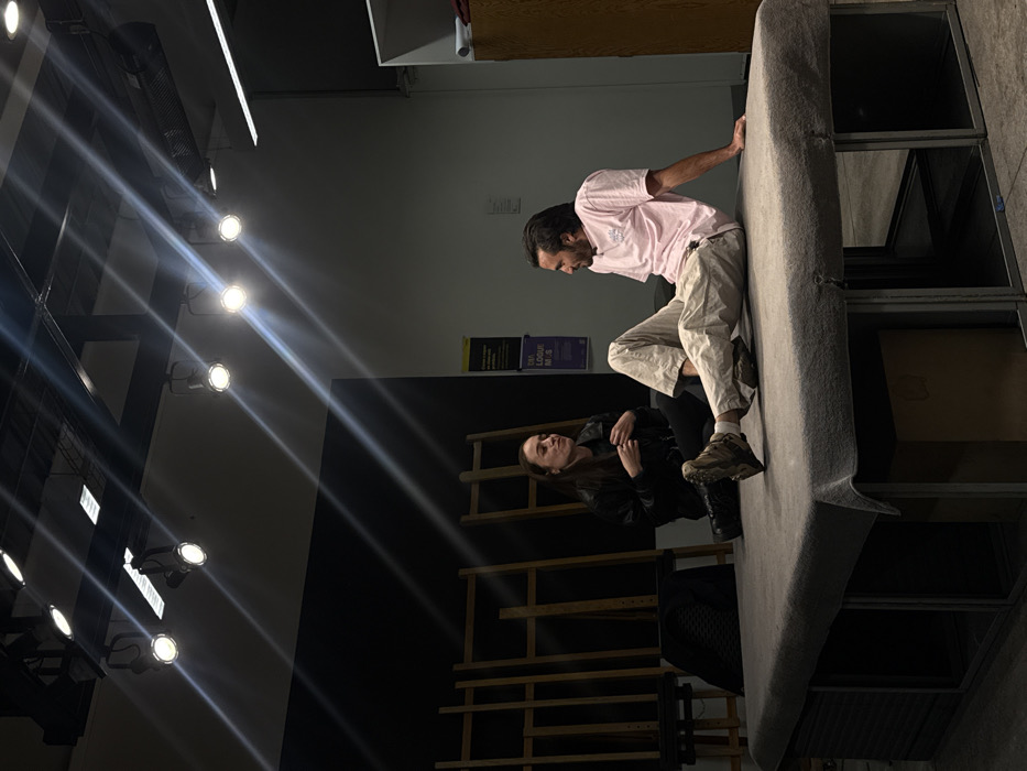 A man in a pink shirt and beige pants leans over the edge of a large, grey upholstered structure, possibly a stage prop. A woman in a black jacket sits below him, seemingly looking up. In the background, there's a black wall with a wooden structure, possibly a set design element. Stage lighting with spotlights illuminates the scene from the upper left corner. Storage racks and other materials are visible to the right.