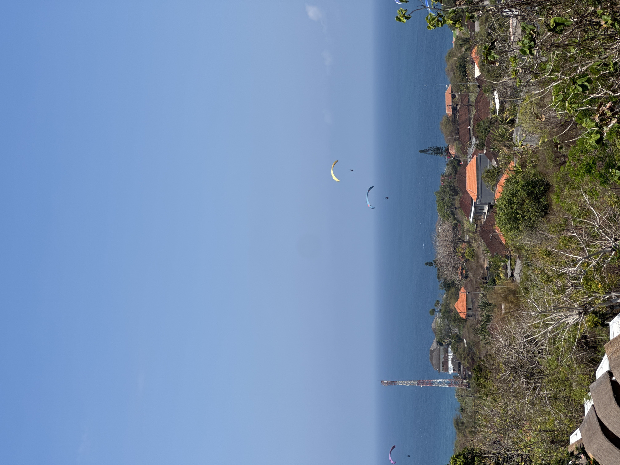 The image depicts a coastal landscape in Kuta Selatan, Indonesia, under a clear blue sky. In the foreground, dense vegetation, including dry, bare trees and green foliage, covers a hilly terrain. A few thatched-roof buildings are partially visible at the bottom left. Further into the scene, a village with terracotta-tiled roofs is nestled on the hillside. A prominent communication tower stands near the coast. The middle ground features the deep blue ocean stretching to the horizon. Several paragliders are visible in the sky, suspended above the water and the village, suggesting an area known for paragliding activities. The overall environment appears to be a relatively undeveloped coastal region with a blend of natural landscape and residential areas.