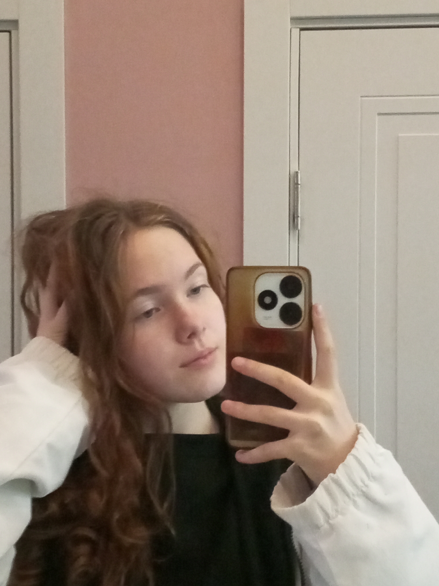 The image displays a mirror selfie of a young individual, likely female, with long, wavy, reddish-brown hair. The subject is wearing a dark-colored top under a light, possibly white or off-white, jacket with a visible elasticated cuff on the right sleeve. Her left hand is raised, touching the hair near the back of her head. Her right hand holds a smartphone, appearing to be an iPhone model with three distinct rear camera lenses, encased in a translucent brown cover. The background consists of an interior space featuring a plain, light pink wall on the left and a white paneled door with a visible silver hinge on the right. The specific location within Saint Petersburg, Russia, cannot be determined from the visual content of the image.