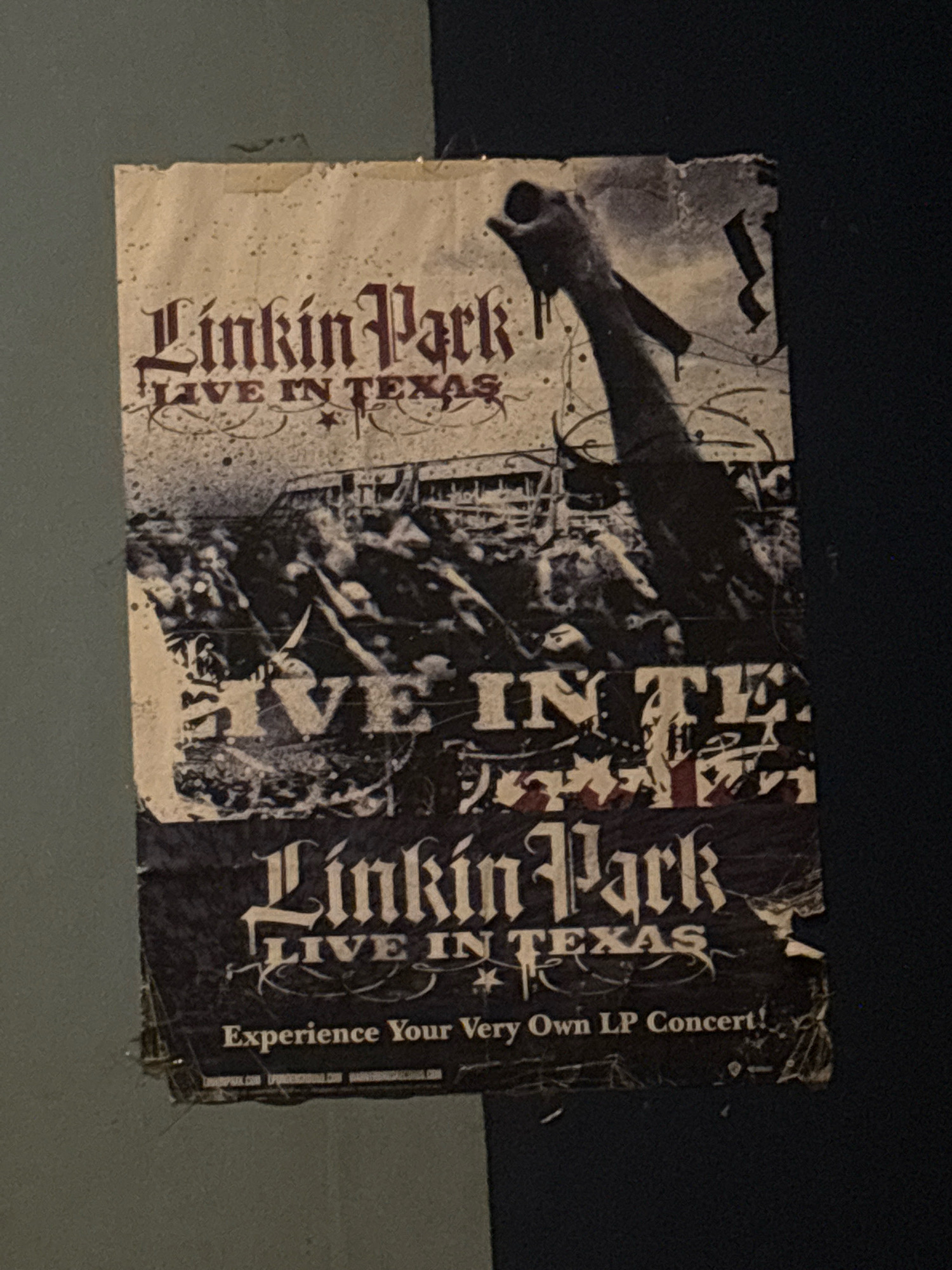 The image is a close-up, slightly angled shot of a poster for "Linkin Park Live in Texas." The poster is weathered and torn, with visible creases and frayed edges, indicating it has been displayed for some time. The poster's design features the band's name in a gothic-style font, followed by "LIVE IN TEXAS." Below this, a dark, gritty image depicts a concert crowd and a stage with a silhouette of a person's arm raised. There are also faint lines and speckles across the poster, adding to its distressed appearance. The bottom portion of the poster reads "Experience Your Very Own LP Concert!" followed by what appear to be website URLs, though they are partially obscured and difficult to read. The poster is affixed to a dark, textured surface, possibly a wall. The lighting is dim, creating shadows and highlighting the worn texture of the poster. There is no information in the image to suggest a location in Bekasi, Indonesia.