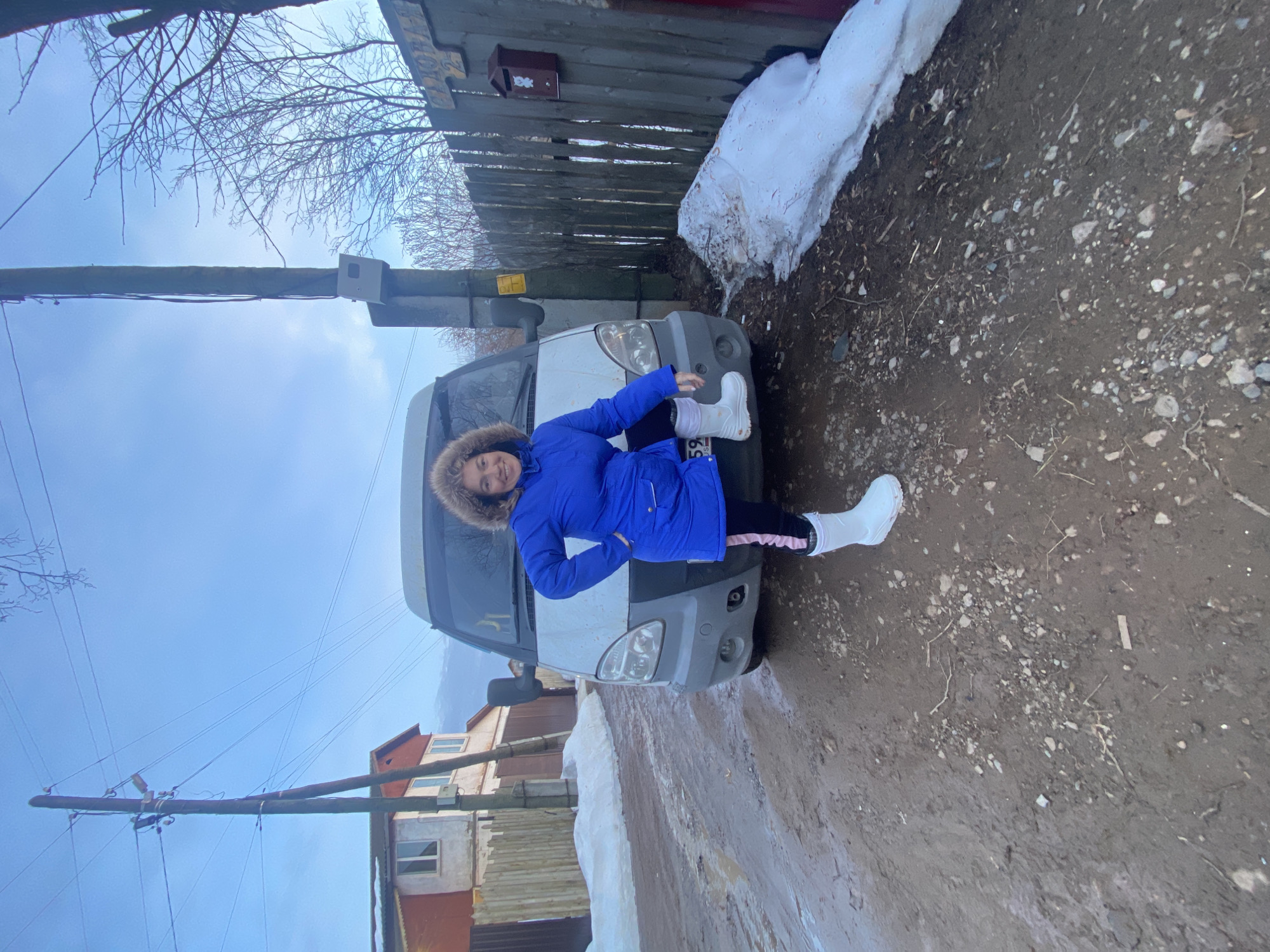 A woman is captured mid-pose on a muddy, unpaved street in Ольховка, Russia. She stands prominently in the foreground, smiling directly at the camera. She has curly brown hair and is dressed in a bright blue hooded parka with a fur-lined hood, black leggings featuring a pink stripe on the left outer seam, and white winter boots. Her right hand rests on her hip, and her left foot is elevated, resting on the front bumper area of a white panel van positioned directly behind her.
The van, possibly a GAZelle model, is partially visible, displaying a light gray lower bumper and headlights. A portion of a Russian license plate, "59 RUS," is discernible on the front bumper. The ground is composed of dark, wet mud mixed with small rocks and debris, indicating unpaved conditions and possibly recent thaw.
To the left, a two-story house with a red roof and light-colored walls is visible behind a wooden picket fence. On the right, a weathered wooden fence runs along the roadside, featuring a small, reddish-brown mailbox adorned with a white animal decal. Above the mailbox, a sign reads "САДОВАЯ 2" (SADOVAYA 2), indicating a street name and house number. Patches of melting snow are present along both sides of the road next to the fences. Bare deciduous trees and bushes are visible in the background, suggesting a cold season. Overhead, a utility pole stands tall on the left, supporting multiple power lines and a small junction box, set against a clear, light blue sky.