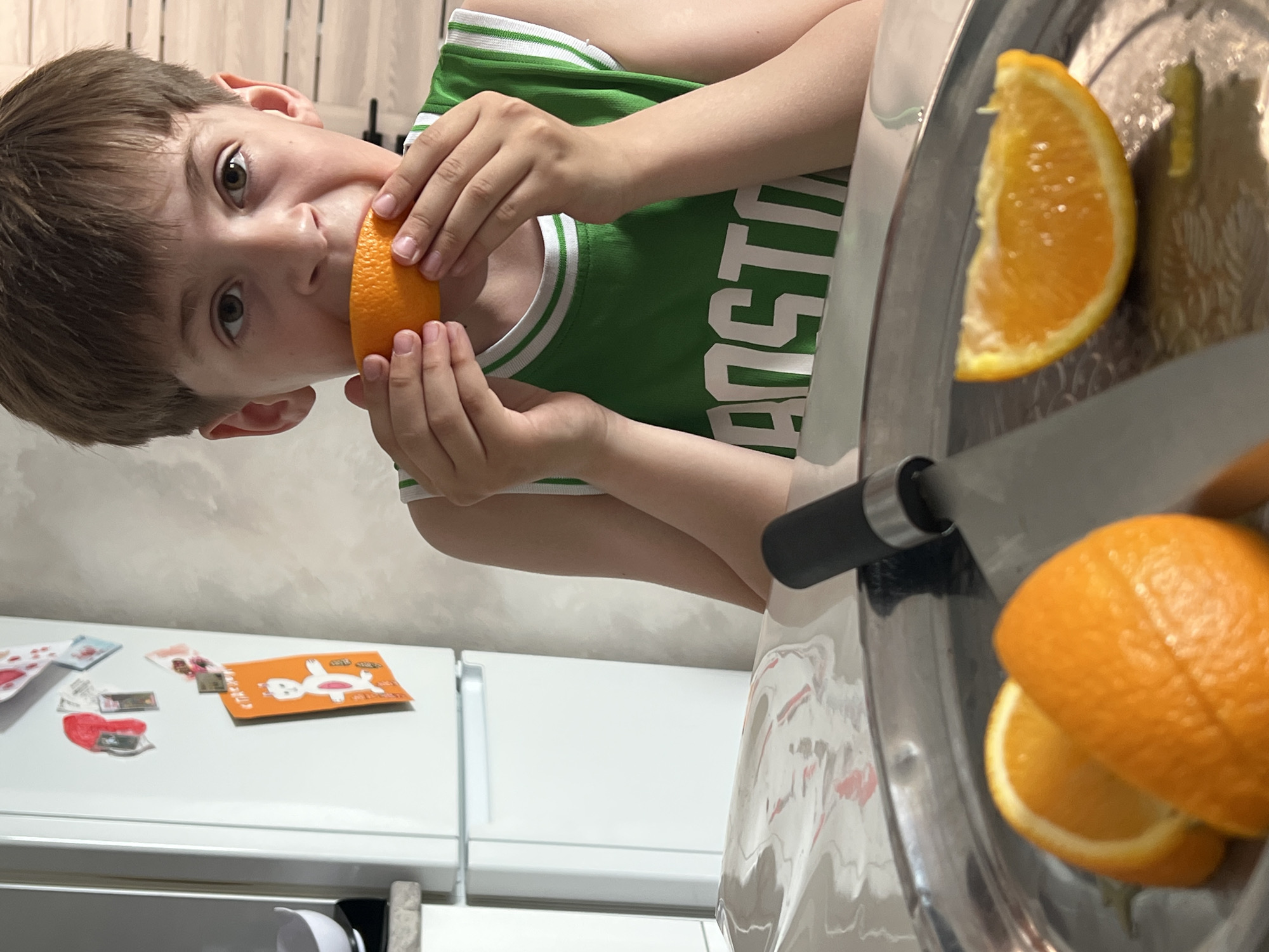 A young boy with brown hair and eyes, wearing a green basketball jersey, is eating a slice of orange. He is holding the orange slice with both hands, bringing it up to his mouth. In the foreground, on a silver tray, are additional orange slices and a knife. In the background, a white refrigerator is visible with magnets and drawings attached. There is no information to suggest this image was taken in Izhevsk, Russia.