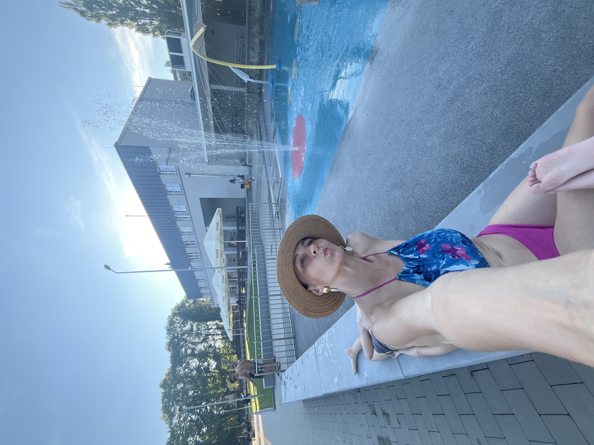 A woman in a straw hat and bikini poses for a selfie on the edge of a splash pad at an outdoor public swimming pool. She is lying on a concrete ledge, facing the camera with her lips pursed. The splash pad features a fountain spraying water into a shallow pool with blue and red sections. In the background, a modern building with large windows and several umbrellas for shade are visible. Other people are seen in the distance, enjoying the pool area. The sky is clear and blue, with the sun shining brightly.