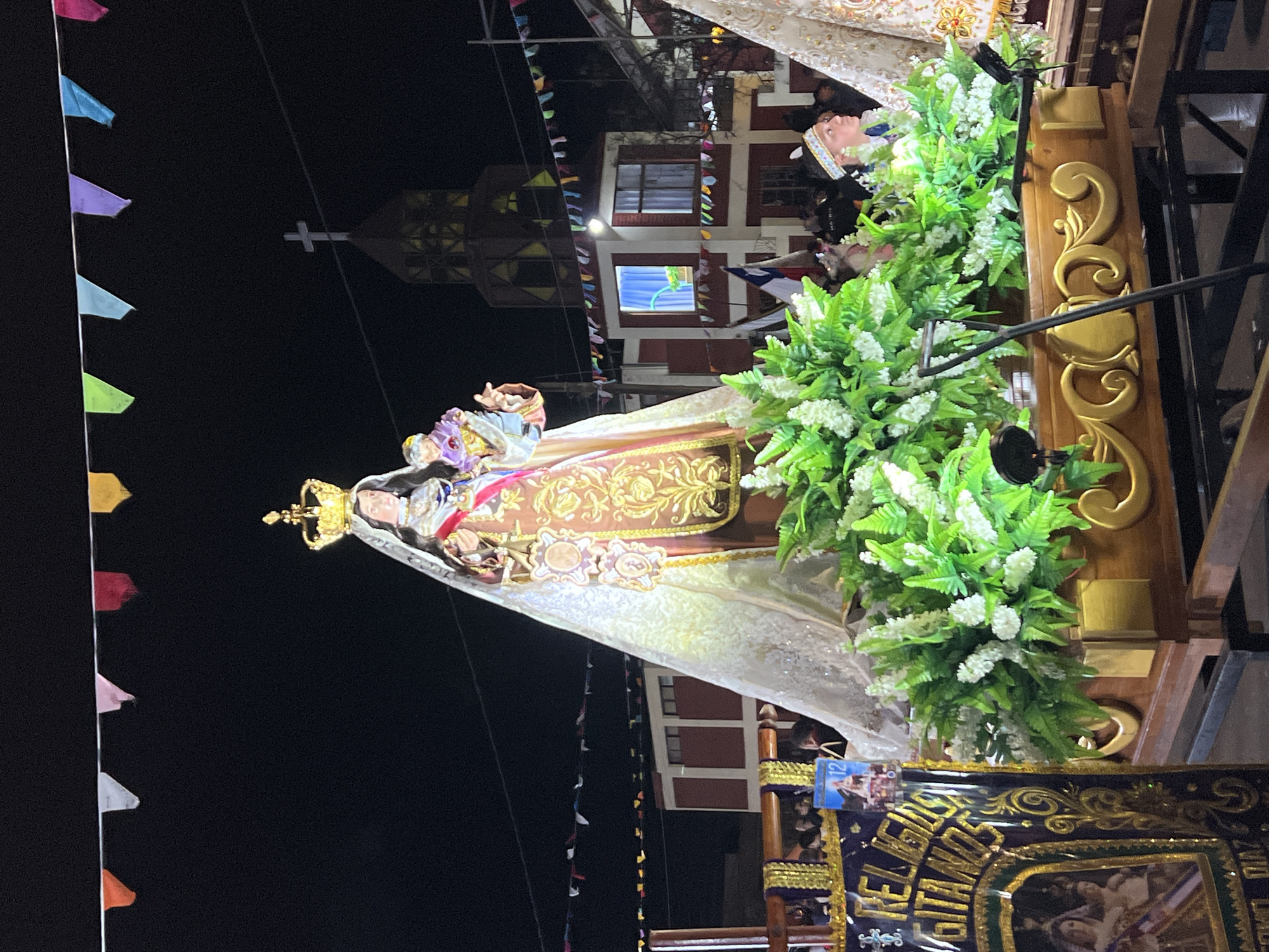 The image depicts a religious procession at night in Arica, Chile. The central focus is a large statue of a crowned woman holding a baby, adorned with a golden and brown robe featuring intricate embroidered patterns. The statue is mounted on a decorated wooden float, surrounded by abundant green foliage and white flowers. Festive triangular flags in various colors are strung across the dark sky above. In the background, a building with a cross atop its tower is visible, along with other structures adorned with flags and lights. Several people are present, some in traditional attire, including one person wearing a feathered headband and another in the foreground looking to the right. A sign with gold lettering that reads "RELIGIOSA GITANOS" is partially visible on the left. The overall scene suggests a religious festival or celebration taking place in the city.