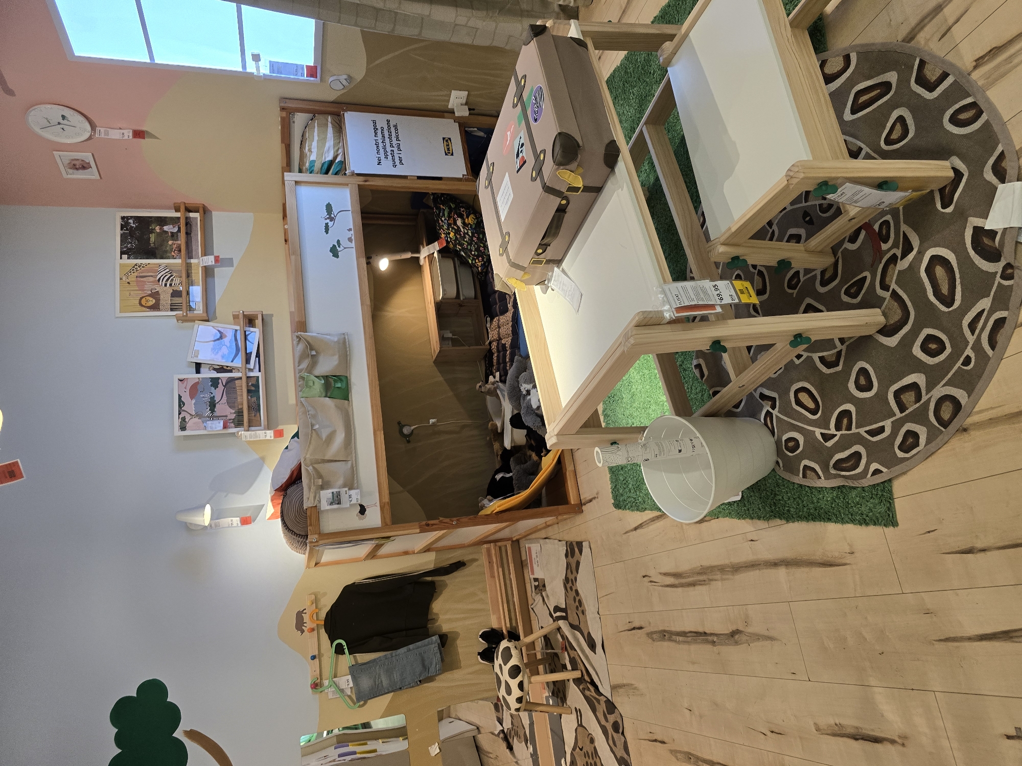 This image captures a playful and organized children's play area, likely within a retail environment like IKEA, as suggested by the price tags and signage. The scene is brightly lit, indicating daytime. The dominant subjects are furniture and decorative items designed for children.
A wooden easel-like structure with white boards is a central piece, positioned on a circular rug with a brown and white animal print pattern. A plush toy suitcase, adorned with various stickers, rests on the easel. Various tags are visible on the furniture. Nearby, a white bucket sits on a patch of artificial green grass.
The background reveals a tiered wooden structure, perhaps a bed or shelving unit, filled with stuffed animals. On the wall to the left, a clock with animal figures is mounted above a framed picture. Further down the wall are shelves displaying children's books and artwork. A white wall-mounted lamp is also visible. On the lower left, a whimsical tree mural is painted on the wall.
Below the wooden structure, a rug with a giraffe pattern is laid out on a light-colored wooden floor. A small, patterned stool sits on this rug, and a pair of blue jeans hangs from a hanger. In the background, through an opening, a glimpse of more stuffed animals and a yellow hammock-like structure can be seen.
The overall impression is one of a cheerful and stimulating environment designed for children's imagination and play. The display appears to be set up in a store in Baronissi, Italy, judging by the location context. Visible text includes price tags and signage in Italian, such as "Nei nostri negozi applichiamo questa protezione per i più piccoli" which translates to "In our stores, we apply this protection for the little ones."