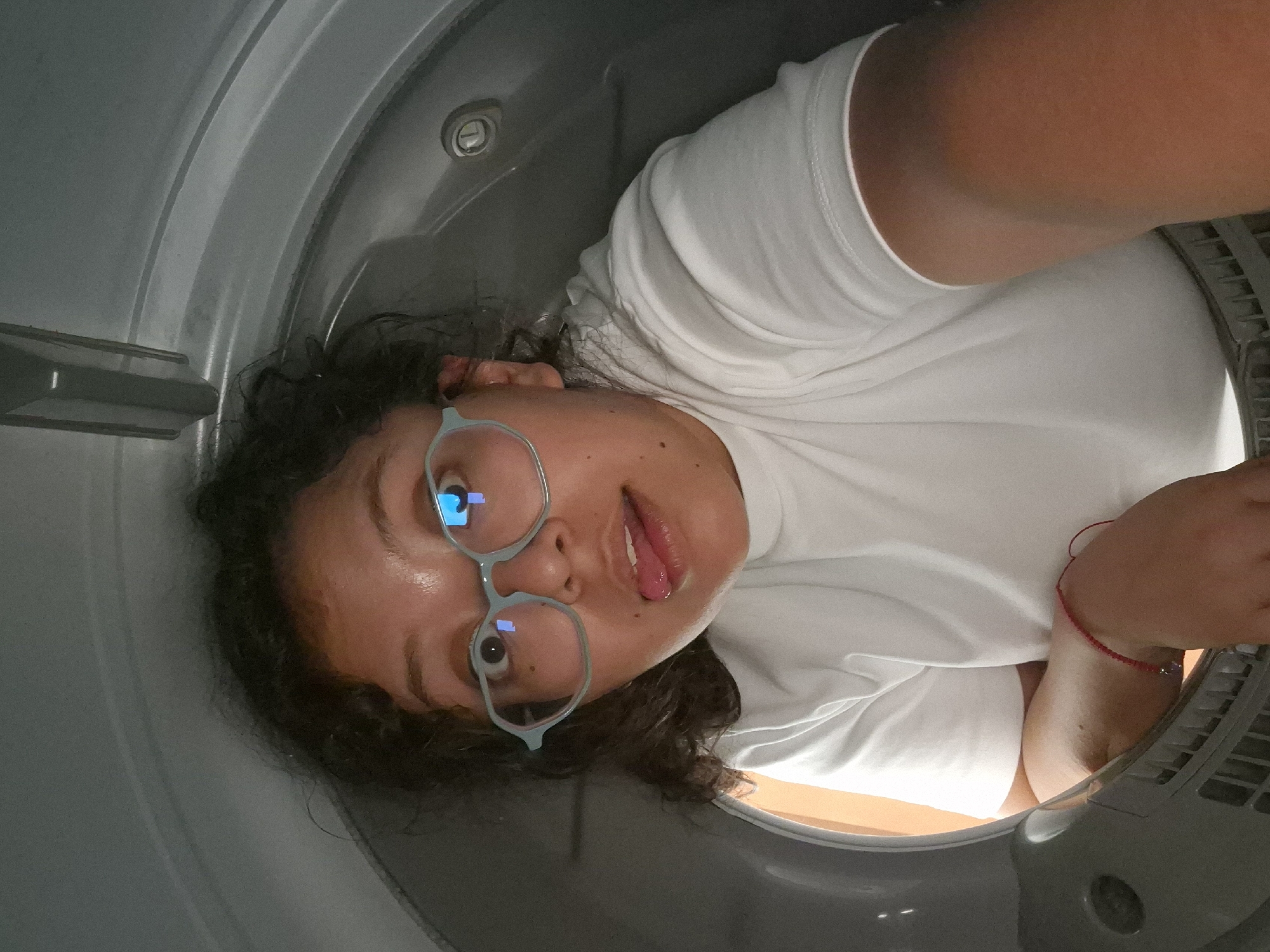 A person wearing glasses and a white t-shirt is inside a dryer. The location is not identifiable from the provided image.