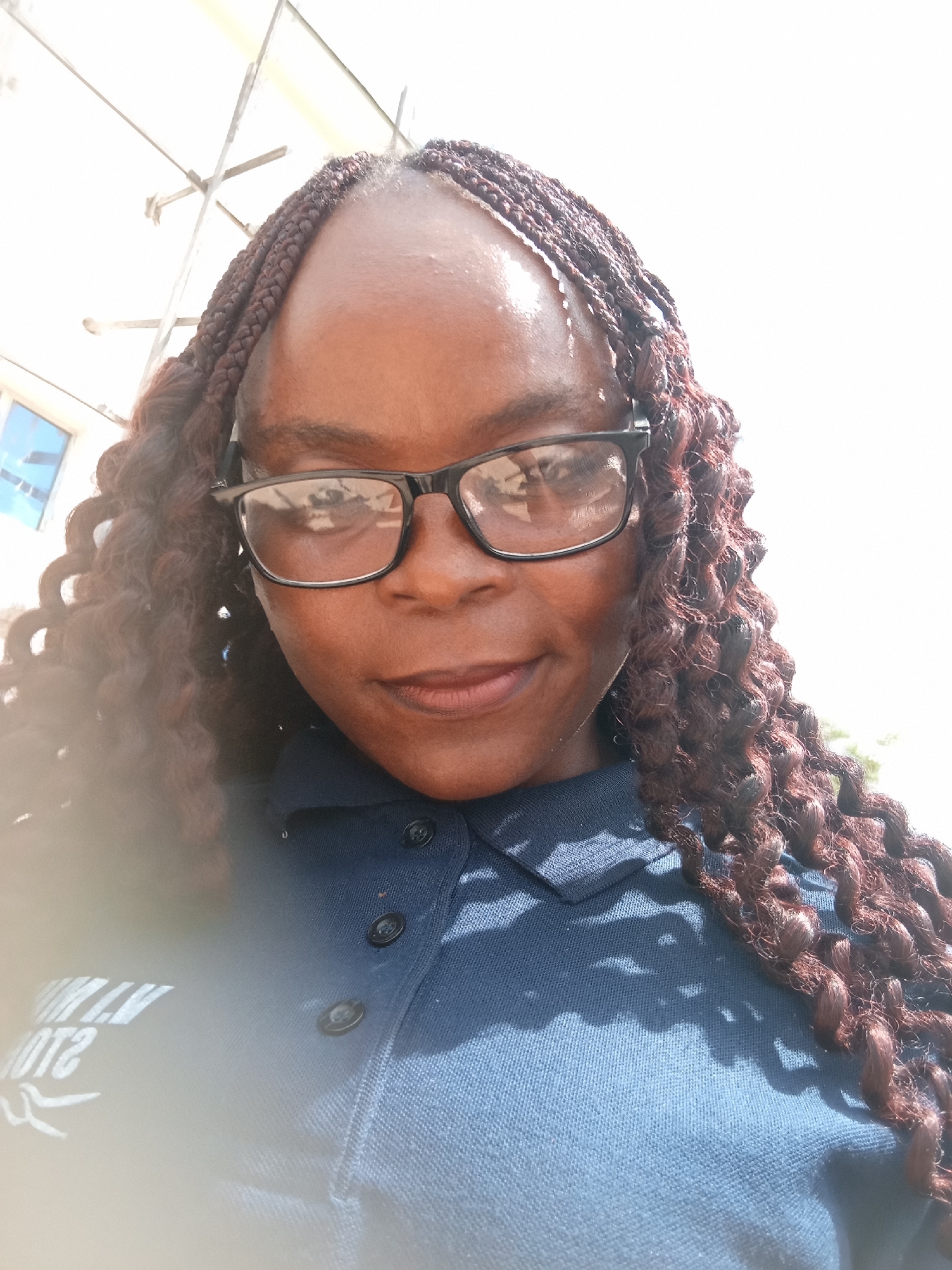 A close-up selfie shows a Black woman wearing glasses and a dark blue polo shirt. Her hair is styled in long, reddish-brown braids. The background is bright and out of focus, with some scaffolding visible in the upper left corner. A hint of a blue-tinted window is also present on the left side. The woman's shirt has white lettering that appears to be a logo, but the text is partially obscured by light reflection. There are dark buttons on the front of her shirt. The scene is outdoors, suggesting it might be a daytime photo taken in Jalingo, Nigeria, though this is not explicitly stated in the image.