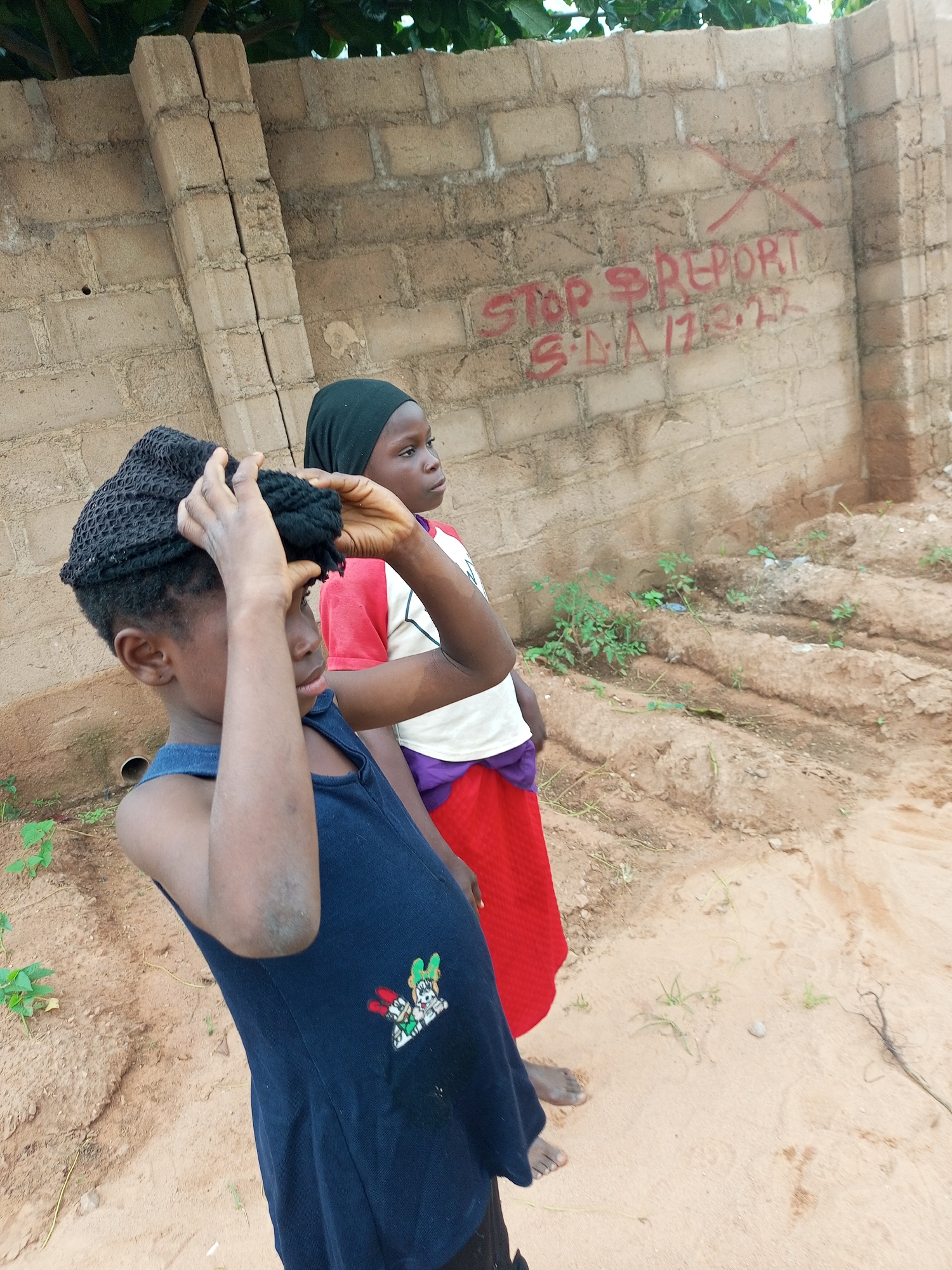 In the city of Tempure, Nigeria, a close-up shot captures two young girls standing in front of a rough, brick wall. The wall has red graffiti on it, reading "STOP REPORT S.I.A 17.3.22." The girl in the foreground, wearing a dark blue sleeveless dress with cartoon characters on it and a black headscarf, is adjusting her headscarf with her hands. The girl behind her, wearing a black headscarf, a white t-shirt with red trim, and a bright red skirt, looks off to the side. The ground is sandy and uneven, with sparse green vegetation growing in patches. The overall scene suggests an outdoor environment, possibly a street or an undeveloped area.