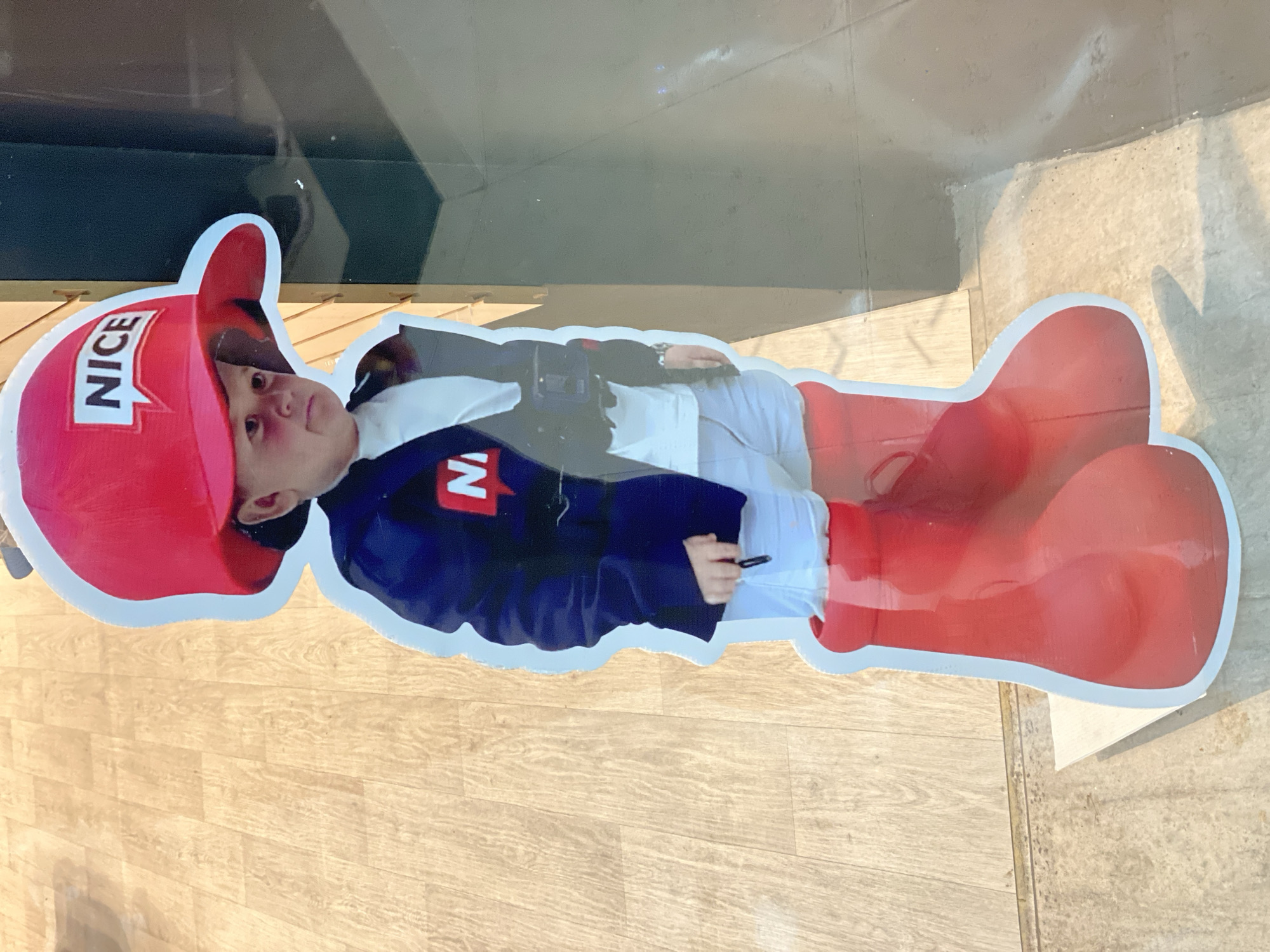 A photograph depicts a cutout advertisement for "NICE" brand goods, featuring a child wearing a red cap with the word "NICE" printed on it and oversized red boots. The child is wearing a dark blue jacket over a white shirt, holding a pen or small object in their hand. The cutout is displayed in a window, with a wooden floor visible below. The image was taken in San Bernardino Tlaxcalancingo, Mexico.
