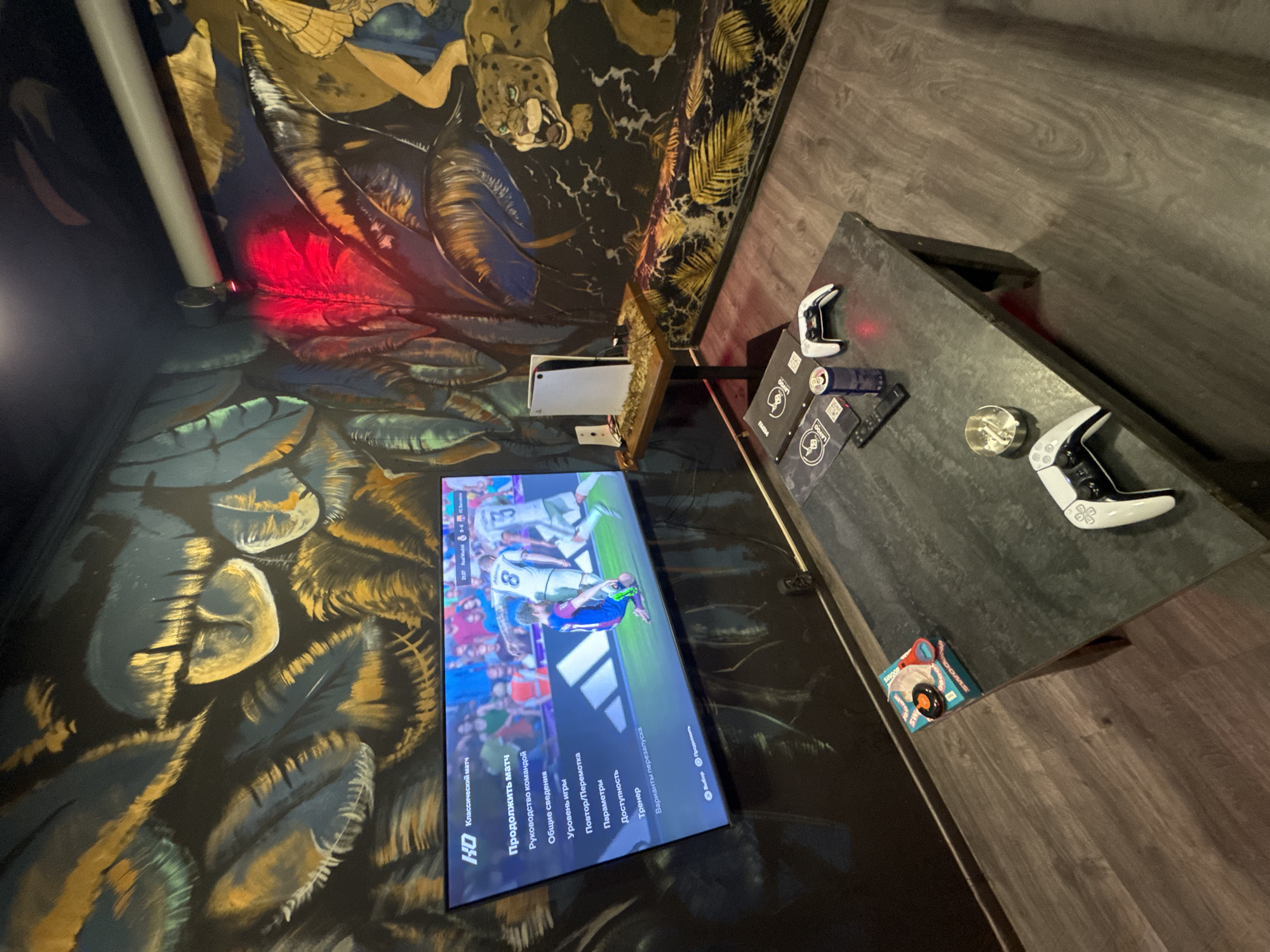 This indoor shot depicts a gaming setup in Yerevan, Armenia, evident from the Cyrillic text displayed on the television screen. The environment is a room with uniquely designed wallpaper: a black background with stylized, gold-accented foliage and an image of a leopard on the ceiling. A white PlayStation 5 console is mounted near the ceiling, connected to a flatscreen TV displaying a soccer game. Below the TV, a gray countertop hosts two white PlayStation controllers and other smaller objects, including a round, metallic ashtray. A box of "SWOOP" brand items is also visible on the countertop. The room appears to have a dim, ambient lighting, with a red light source near the ceiling on the left side of the frame. The overall scene is angled, giving a perspective from a higher vantage point.