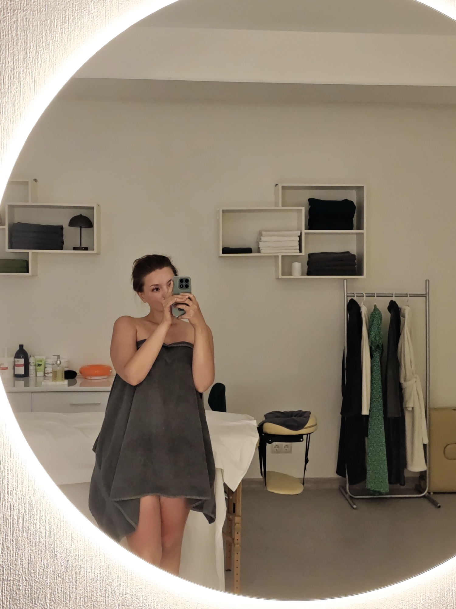 A young woman stands in front of a large, circular mirror, taking a selfie with her phone. She is wrapped in a dark grey towel and appears to be in a spa or treatment room. The room is clean and organized, with white walls and shelves containing neatly folded towels and a small lamp. A rack of clothes, including robes and dresses, stands to the right of the woman. A massage table is visible behind her. The mirror has a bright, illuminated edge. The overall impression is one of a tranquil and professional setting, likely a spa or beauty salon, located in Vinnytsia, Ukraine.