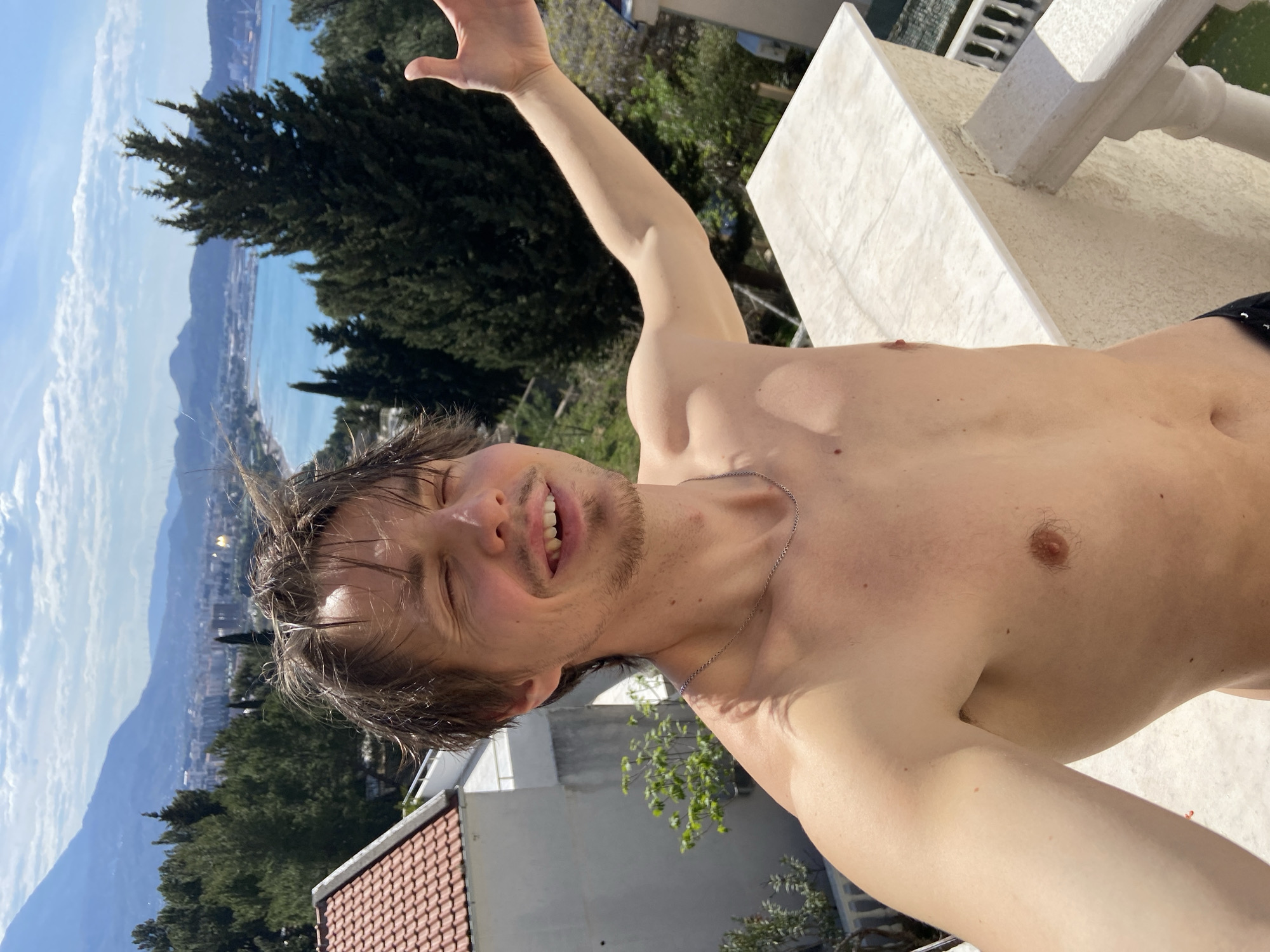 A shirtless man with wet hair is photographed from a low angle. He is fair-skinned with brown hair and a short beard. He is smiling with his eyes closed and has his right arm raised. He wears a thin silver chain around his neck.
In the background, there is a blurred view of the city of Šušanj, Montenegro, along the coast. Buildings with red-tiled roofs and white walls are visible. Green trees are scattered throughout the landscape. Part of a white marble structure is also in the background.