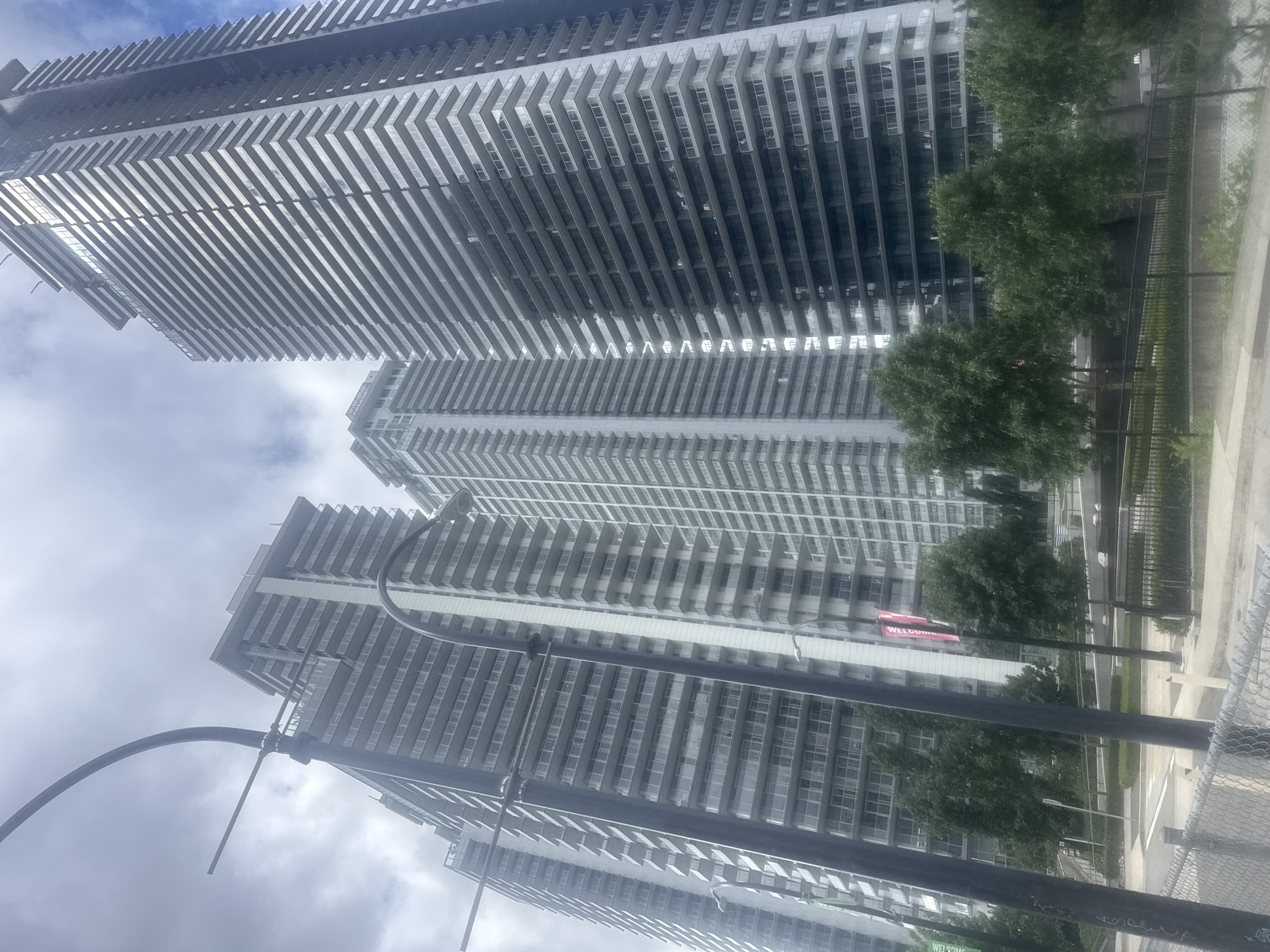 The image captures a view of several tall, modern residential buildings in Burnaby, Canada. The buildings are characterized by their numerous balconies and a contemporary architectural style, with clean lines and a predominantly gray and white color palette. The sky above is overcast, suggesting a cloudy day. In the foreground, there are trees and some street furniture, including lamp posts and a chain-link fence. The overall scene depicts an urban residential area.