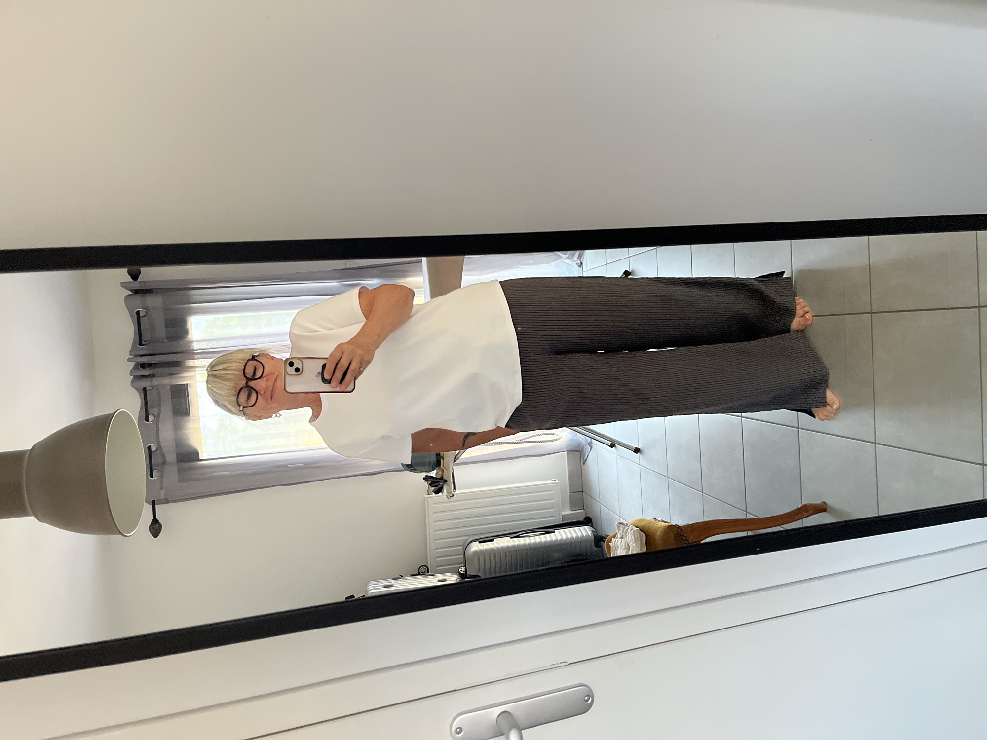 A woman is taking a mirror selfie in what appears to be a home environment, possibly a bedroom or dressing room. She is standing in front of a full-length mirror, reflecting her outfit and the room behind her. She is wearing a white t-shirt and dark, wide-legged pleated pants. Her left hand is in her pocket, and she holds her phone in her right hand to take the picture. She is wearing glasses. In the reflection, we can see a large window with gray curtains, a radiator, an ironing board with an iron on it, a luggage bag, and a small ornate stool with a cushion. The floor is tiled. The overall impression is a casual, domestic setting. There is no visual information in the image that places the scene in Chambœuf, France.