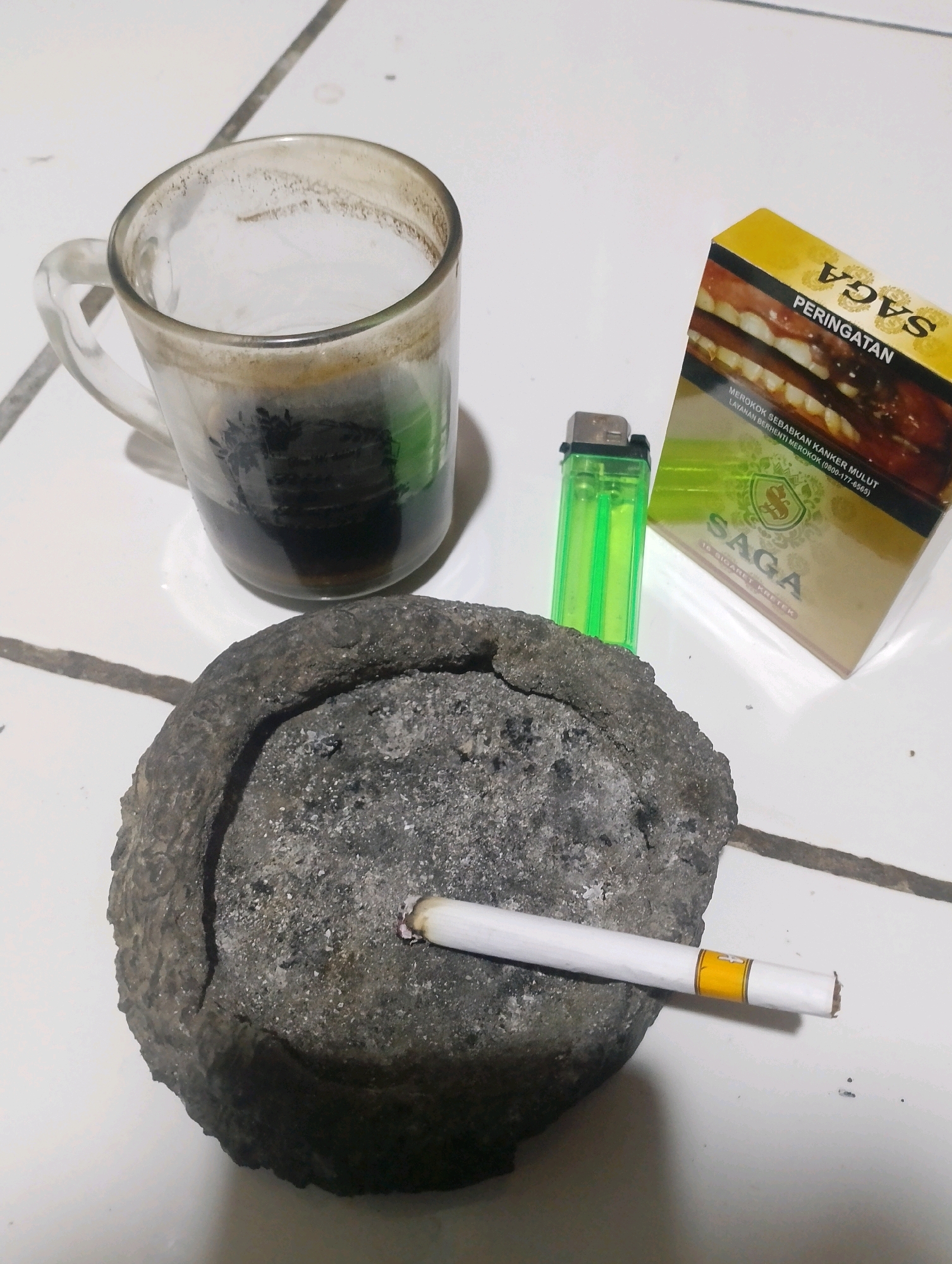 A cigarette rests in an ashtray made of rough, gray stone. The ashtray is filled with ash and cigarette butts. To the left of the ashtray is a clear glass mug filled with dark liquid, possibly coffee, and with residue around the rim. In the background, to the right of the mug, is a pack of "SAGA" brand cigarettes and a green lighter. The objects are placed on a tiled surface with white grout lines. The scene is indoors and appears to be a casual setting, likely a home. The lighting suggests it is nighttime or the room is dimly lit. No people are visible.