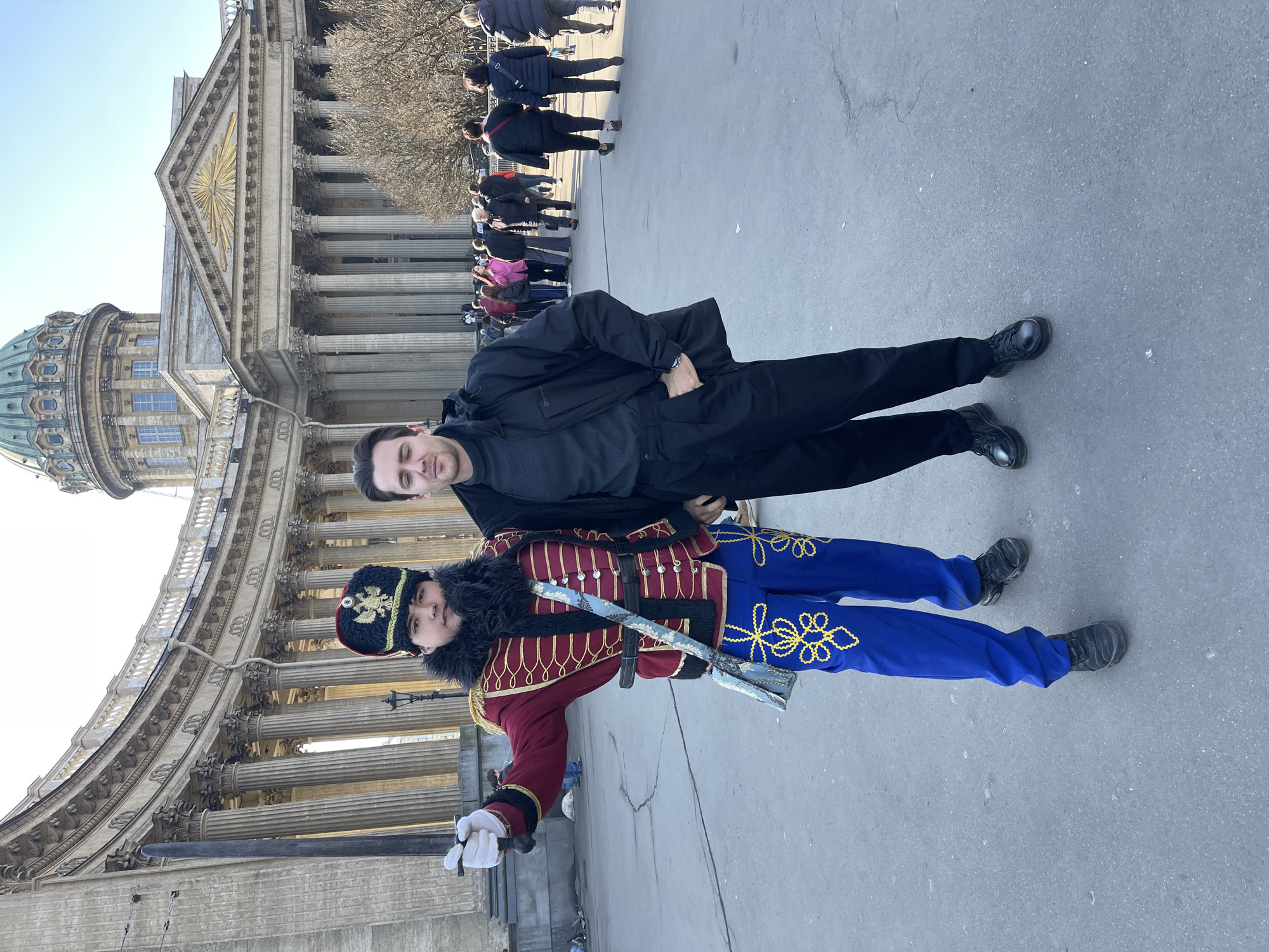 A man in a black jacket poses next to a man dressed in a historical military costume in Saint Petersburg, Russia. The man in costume wears blue pants, a red jacket, and a fur hat. They stand in front of a large building with pillars, likely the Kazan Cathedral. A crowd of people is in the background.