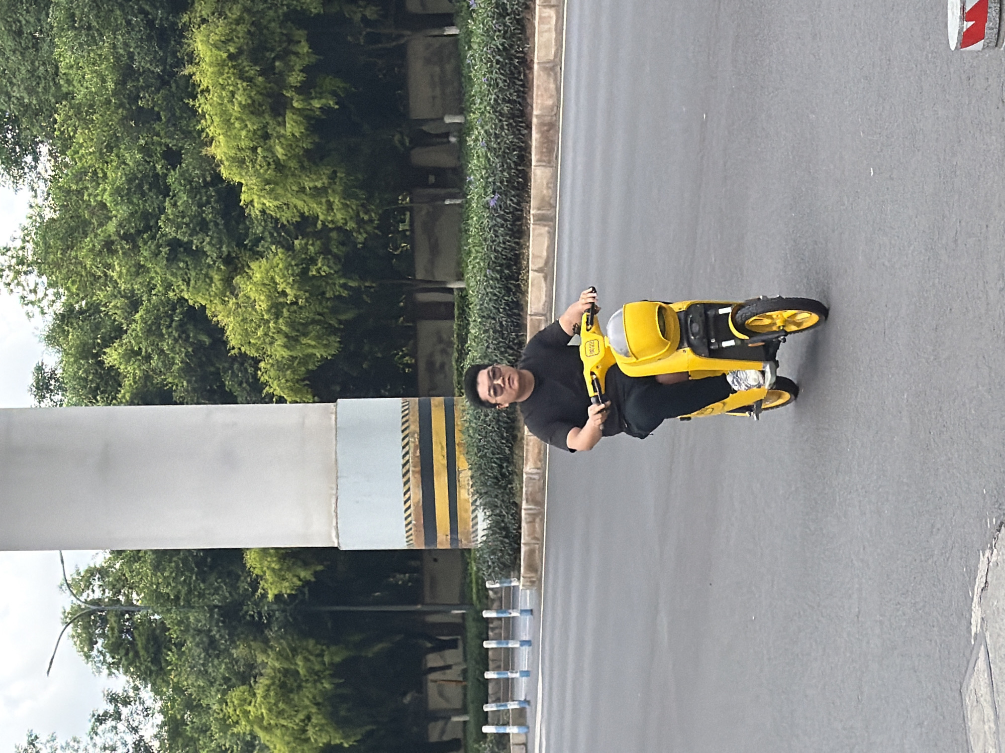 A man is riding a yellow electric scooter on a gray asphalt road. The location is the city of Chongqing High-tech Zone, China. The scooter is oriented horizontally, moving from left to right. The man is wearing a black t-shirt and black pants. Trees and bushes line the side of the road, with a striped barrier adjacent to the foliage. A white pole with black and yellow stripes is visible on the left side.