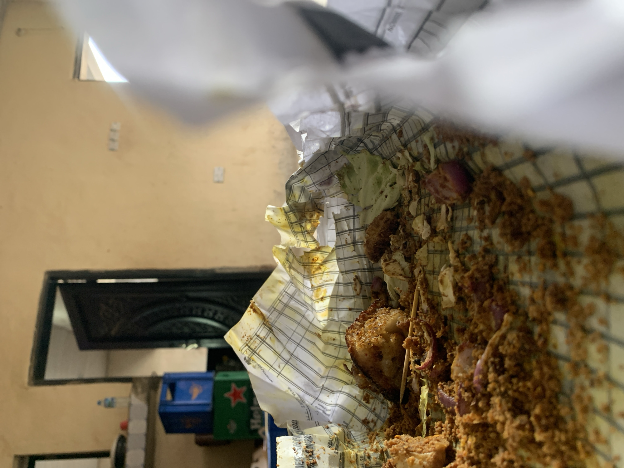 The image appears to be taken inside a building in Kabong, Nigeria. An open door is visible in the background. The main subject is a paper bag containing food, possibly a snack or meal. The food appears to be fried and contains lettuce and red onions. The bag has a checkered pattern. A blue and green object with a red star is partially visible below the bag. A small bottle of water is visible next to it. The surrounding wall is painted a yellowish-tan color and has a window opening.