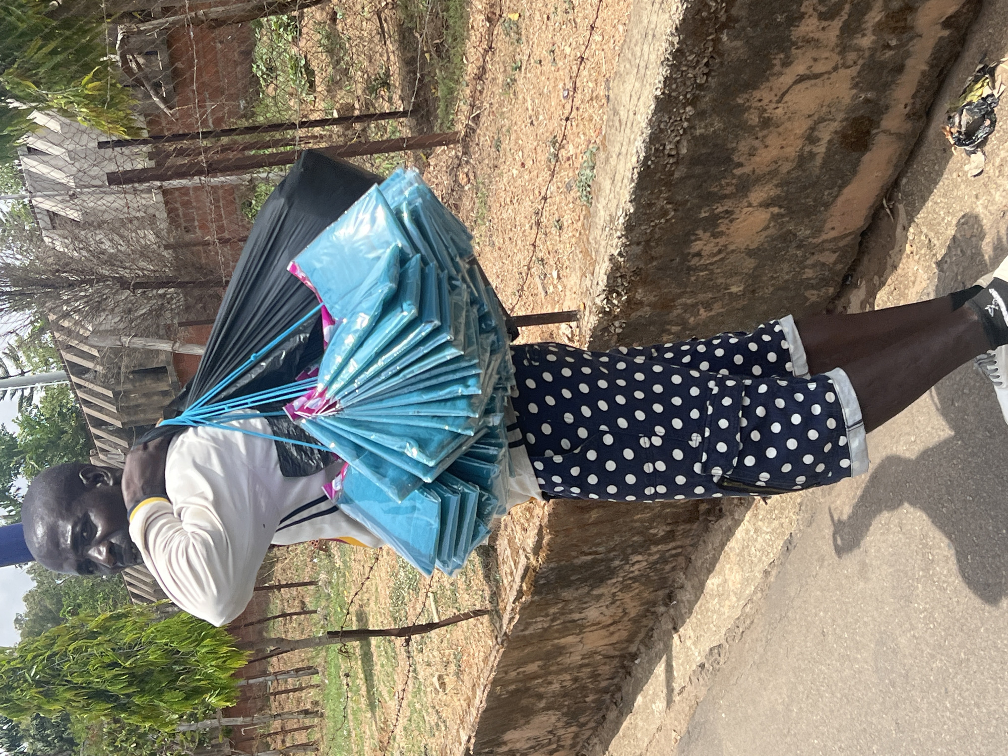 A dark-skinned man is depicted outdoors, likely in Jos, Nigeria, appearing as a street vendor carrying his merchandise. He wears a white long-sleeved shirt with yellow and black accents, and navy blue shorts with white polka dots. He is also wearing white sneakers with dark markings, possibly a logo, on the sides.
The man is laden with goods: a large, dark-colored bundle is strapped to his back, while in front of him, a considerable stack of individually wrapped, light blue, rectangular packages are suspended from his neck and shoulders by blue straps. A small portion of pink material is visible within these items.
The scene is brightly illuminated by sunlight, casting distinct shadows, indicating a clear, sunny day, likely during the mid-day to afternoon hours. The man stands on a paved or concrete surface next to a raised concrete curb. To his left, a dirt area with sparse vegetation and a metal fence (appearing to be chain-link or barbed wire) runs alongside, with brick buildings and lush green foliage visible in the background. The man looks towards the viewer with a neutral expression. No significant text is visible apart from a small, indistinct logo on his shoe.