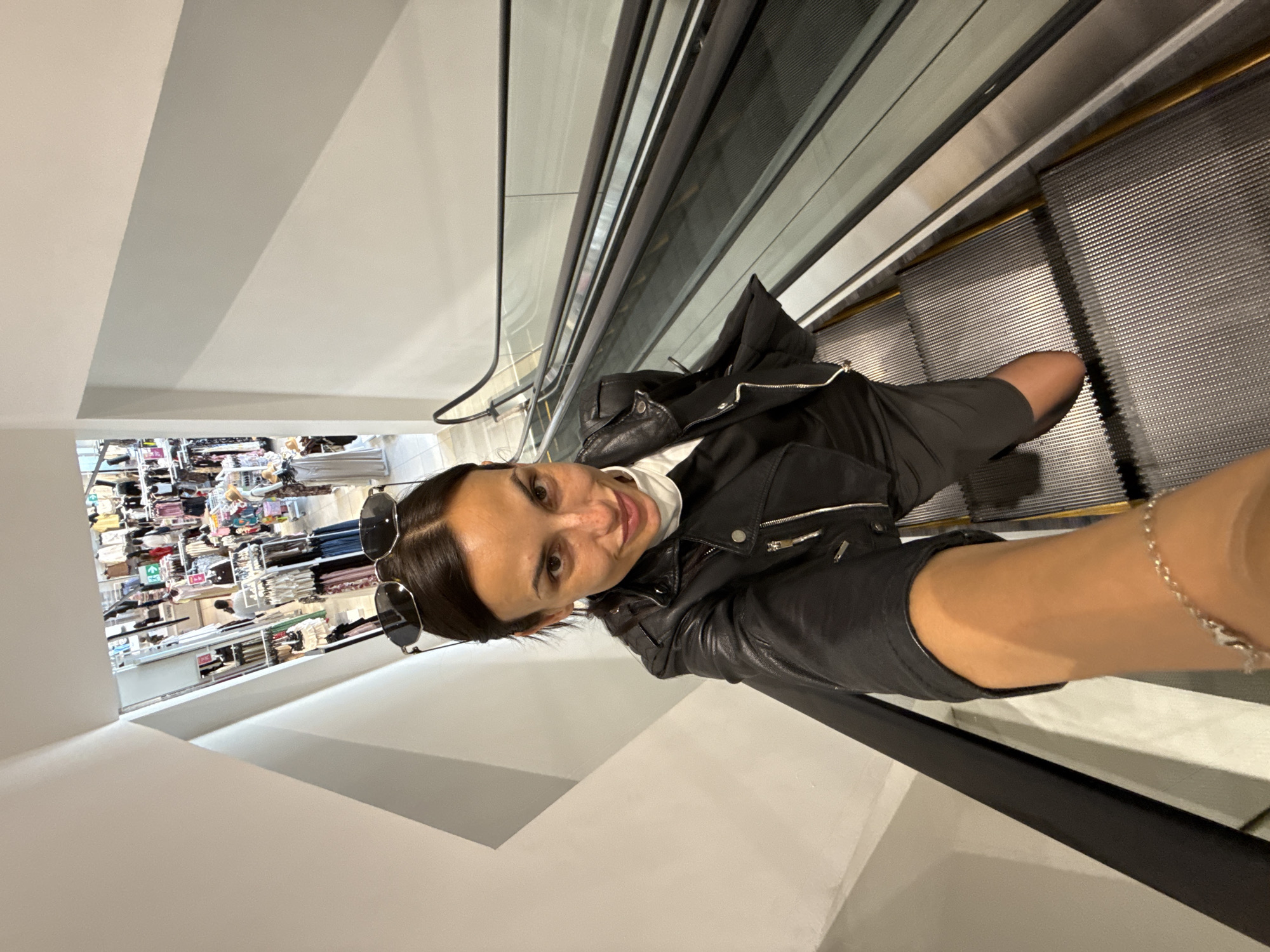 The image shows a woman taking a selfie while riding an escalator. She is wearing a black leather jacket, a white turtleneck shirt, and black shorts. Sunglasses are perched on her head. The escalator appears to be inside a retail environment, as a clothing store is visible in the background. The location is potentially a shopping center in the city of Pleven, Bulgaria, although this cannot be definitively determined from the image alone.