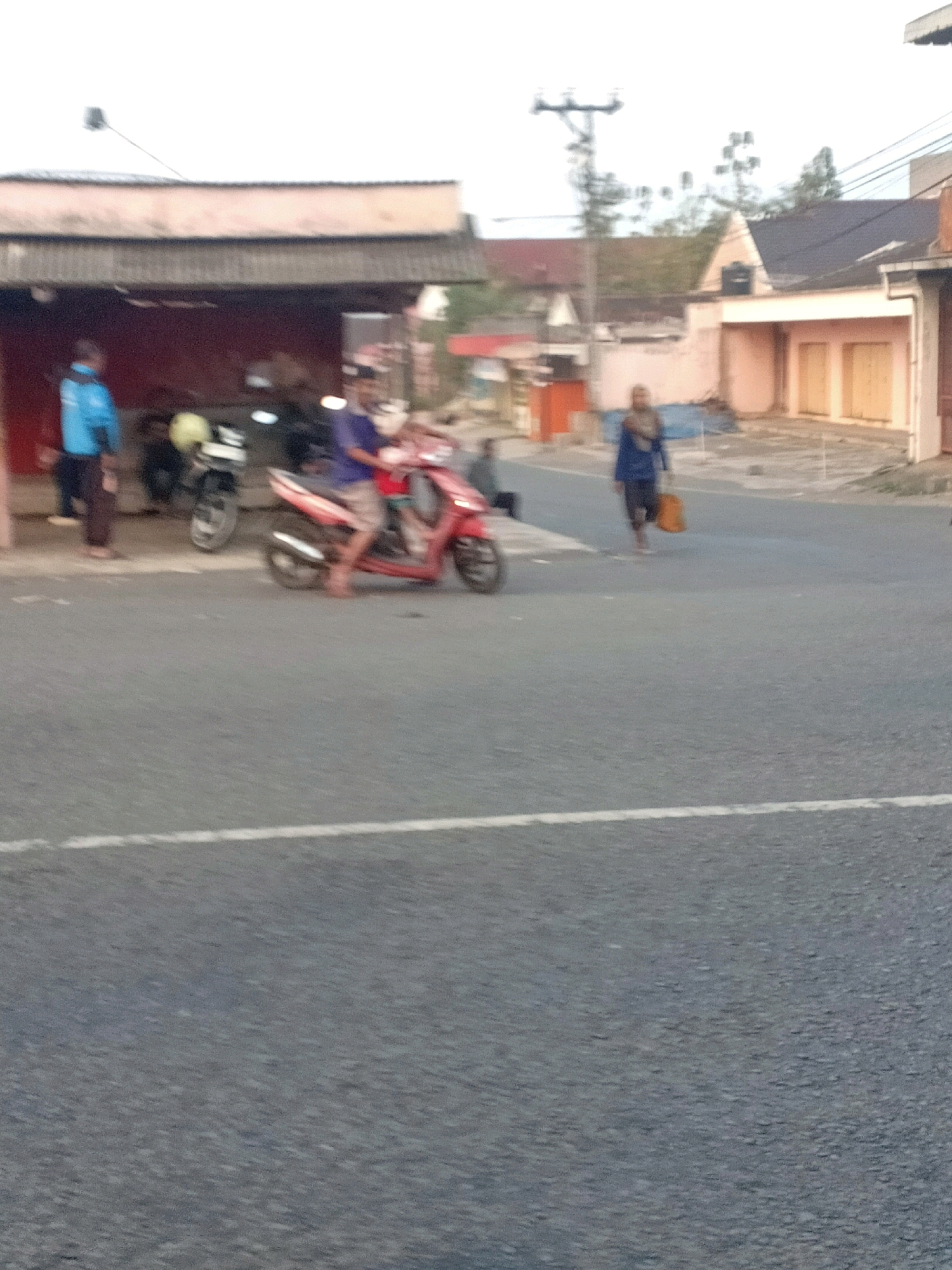 The image depicts a street scene in Mojoreno, Indonesia. In the foreground, a red scooter is parked on the asphalt road. A person is seated on the scooter, wearing a dark blue t-shirt and light-colored shorts. To the left of the scooter, another person in a bright blue jacket and dark pants stands near a building with a dark roof and a pink facade. Behind them, a motorcycle with a yellow seat is visible. Further down the street, a person in a brown headscarf and dark clothing walks away from the viewer, carrying an orange bag. Buildings with light-colored walls and cream-colored doors line the street. The sky is overcast.