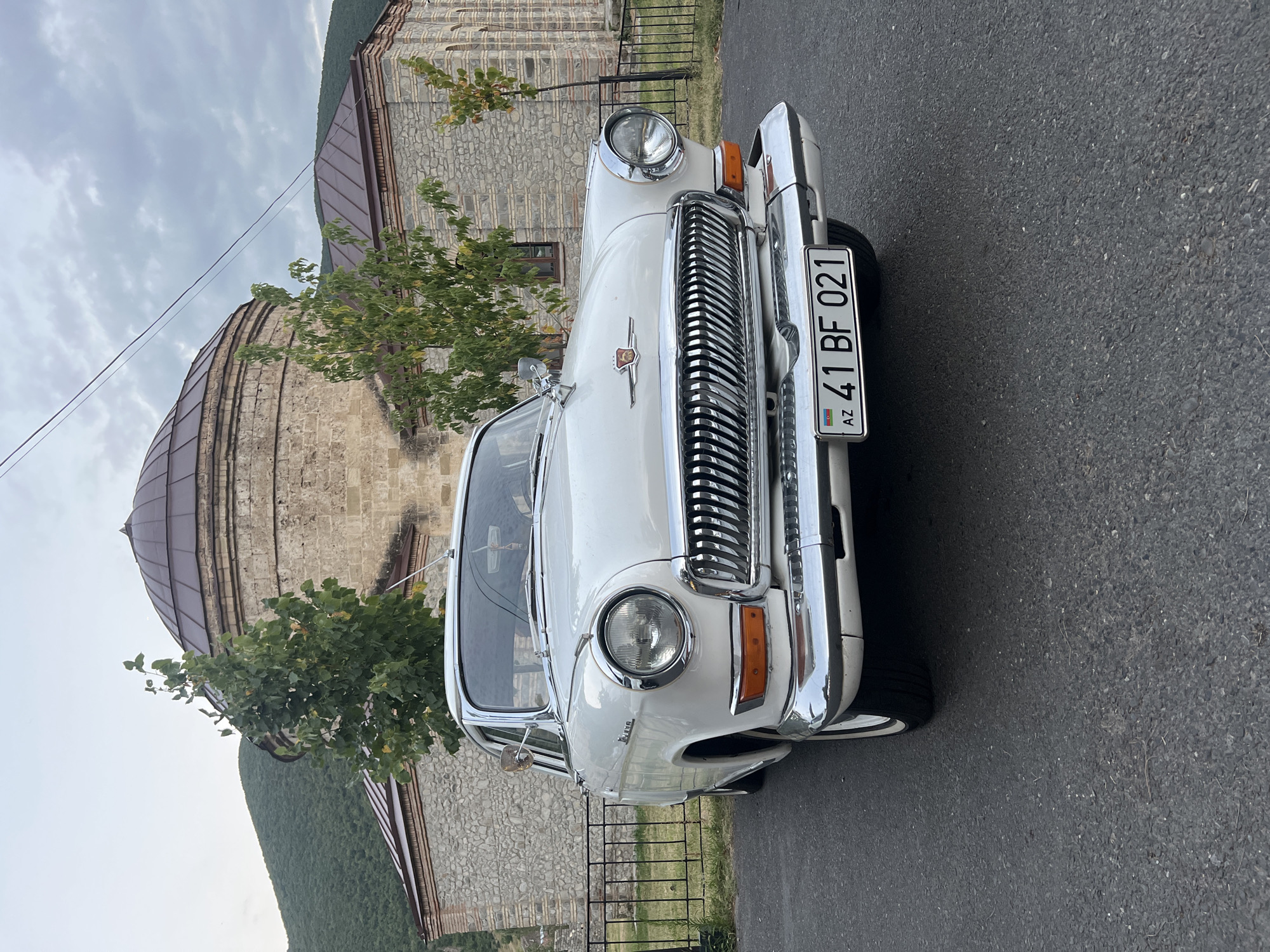 A white, vintage Soviet-era car, identified by its license plate as AZ 41 BF 021, is parked on an asphalt road in front of a stone building with a domed roof. The building appears to be a historical site, possibly a mosque or caravanserai, located in the city of Shaki, Azerbaijan. The car is angled towards the viewer, showcasing its chrome grille, circular headlights, and a red emblem on the hood. Trees with green foliage are visible behind the car, and a green mountain range forms the backdrop under a cloudy sky. A black metal fence separates the car from the building. The scene is captured from a low angle, emphasizing the car's presence.