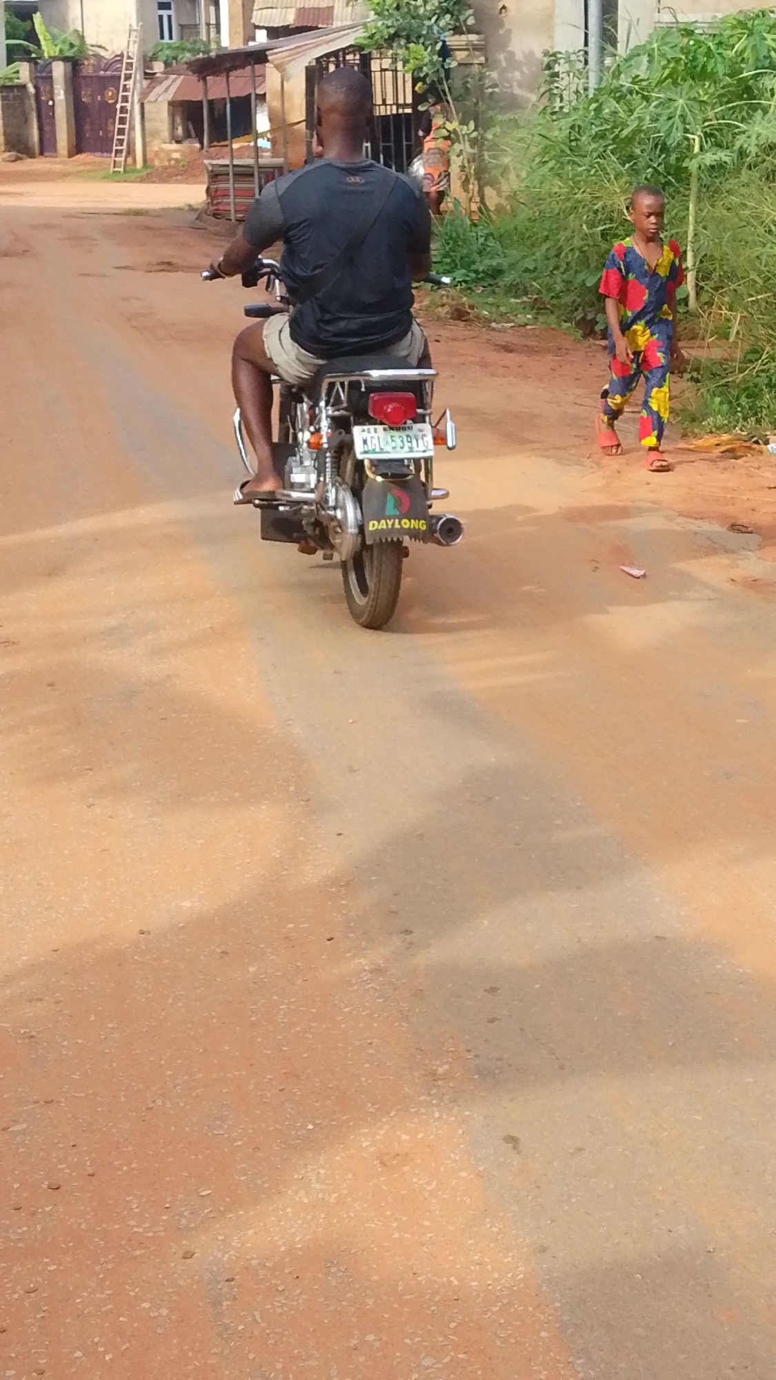 The photo shows a man riding a Daylong brand motorcycle away from the camera on a dirt road in what appears to be a residential area of Enugwu-Ukwu, Nigeria. The license plate on the motorcycle reads MGL-539YG. A child wearing a colorful jumpsuit is walking along the side of the road to the right of the motorcycle. Houses, a metal gate, and vegetation are visible in the background.