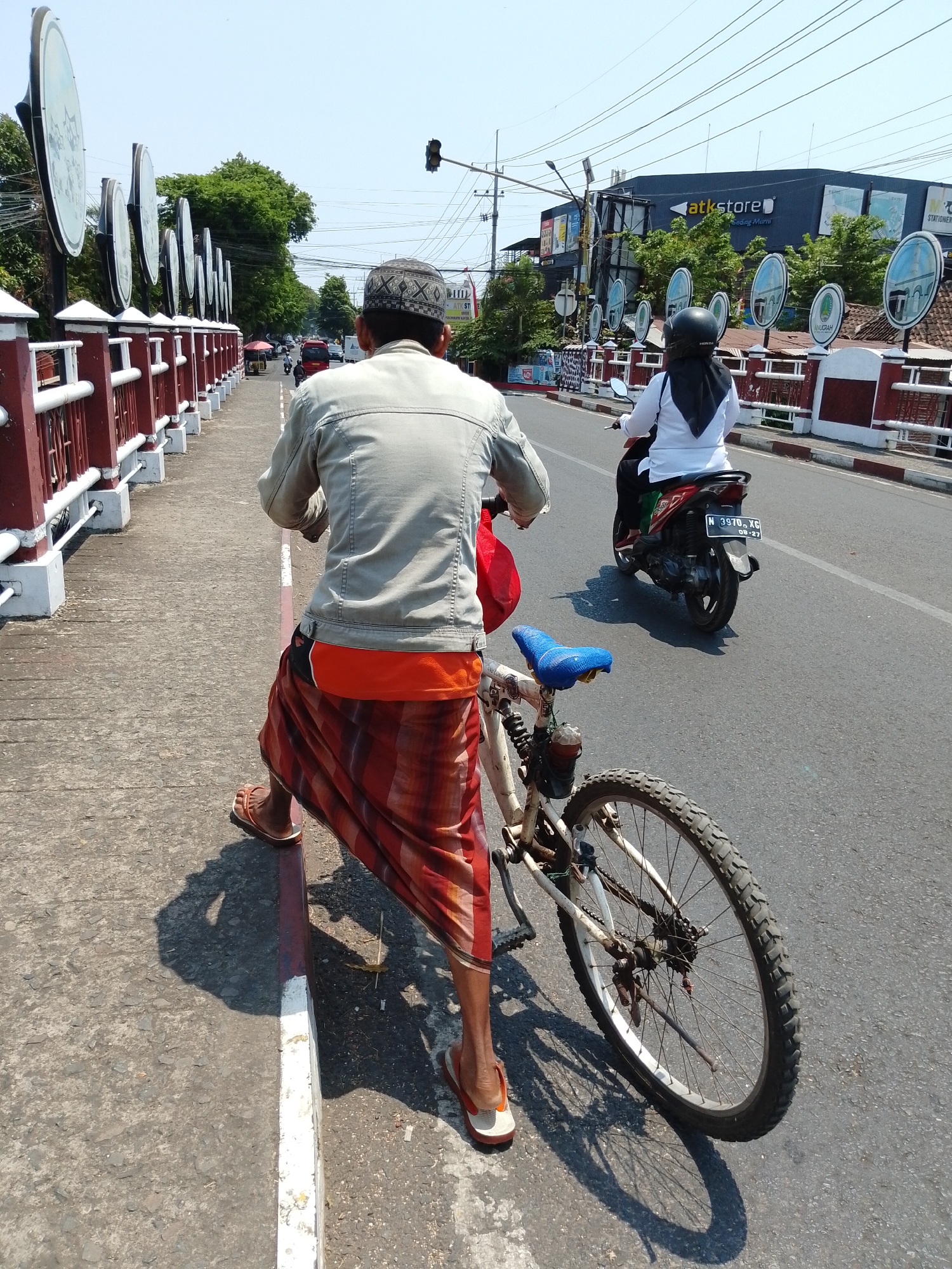 The image shows a man on a bicycle on a sidewalk in Pasuruan, Indonesia, with a scooter on the road behind him. The man is wearing a light-colored jacket, an orange shirt, and a red and orange checkered sarong. He is also wearing sandals and a patterned cap. The bicycle is white with a blue seat and has a red bag attached to the handlebars. The scooter is red and black, and the rider is wearing a white shirt and black headscarf. In the background, there is a building with the word "atkstore" visible, trees, traffic lights, and power lines. The sidewalk has a red and white border, and the road is asphalt. The sky is clear and blue.
